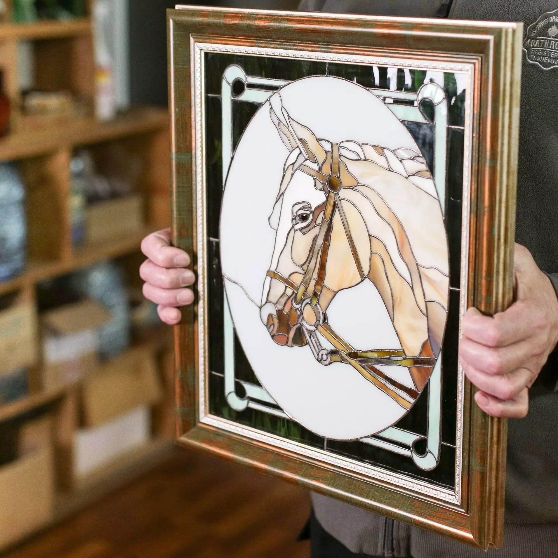 Person holding a framed stained glass panel with a beige horse portrait and a green-bordered frame.
