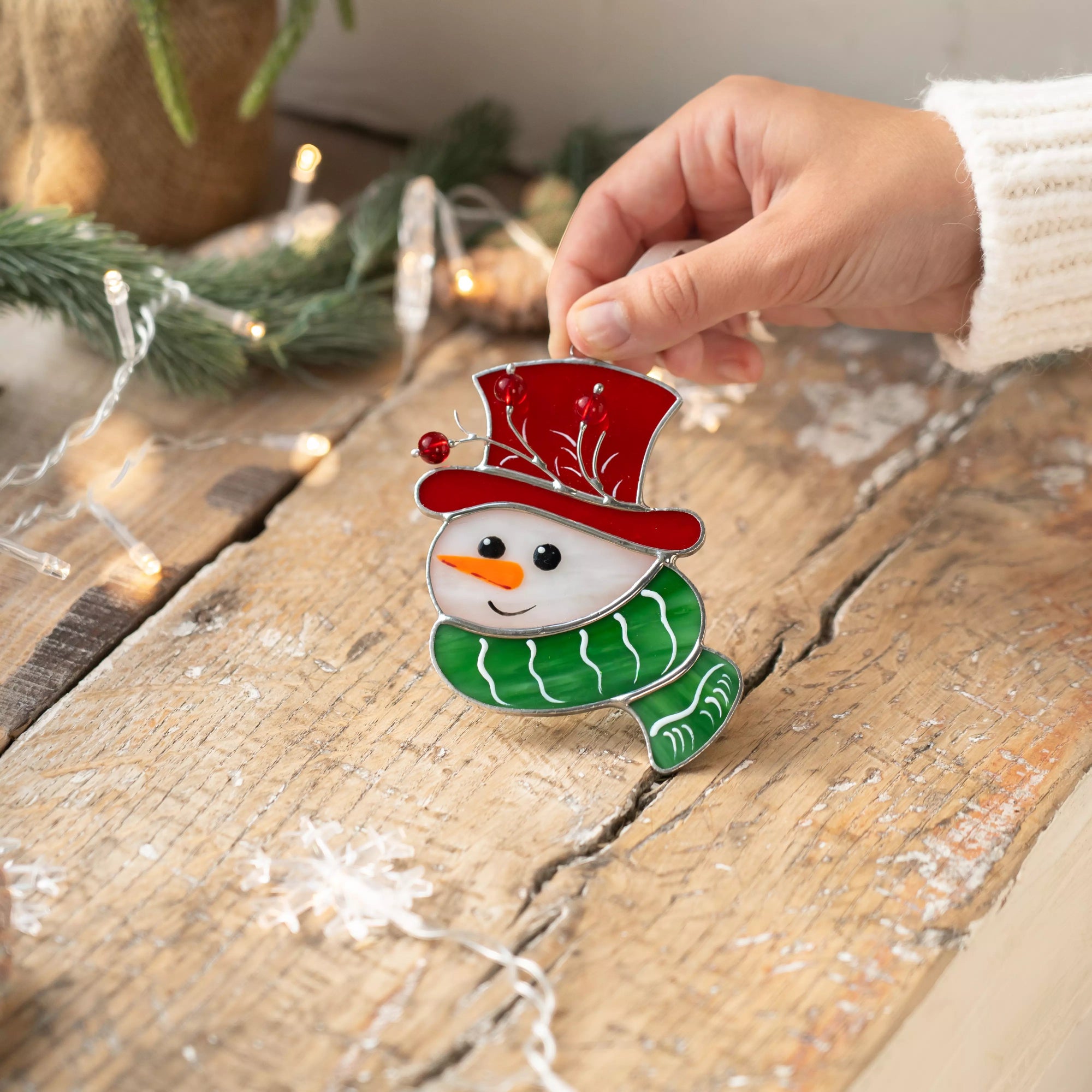 Stained glass snowman with red hat and green scarf resting on a rustic wooden table surrounded by lights.