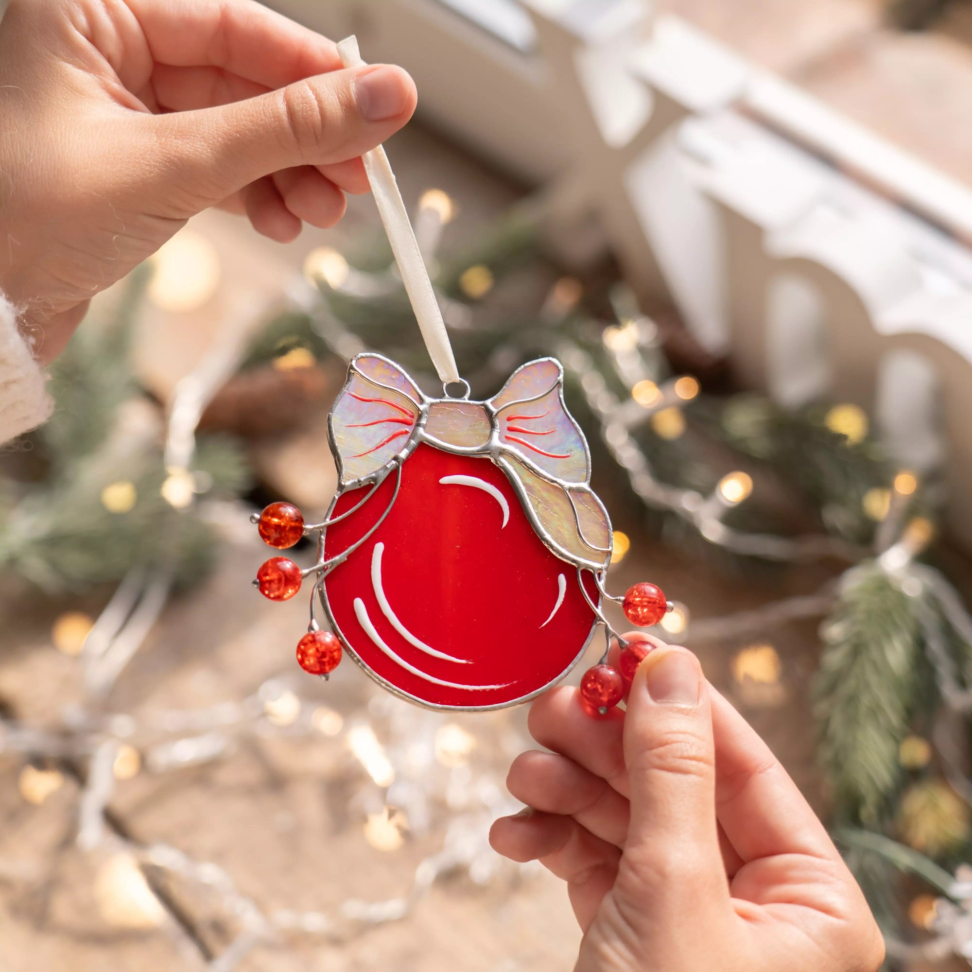 Hands holding a red stained glass Christmas ornament with a bow against a festive background.