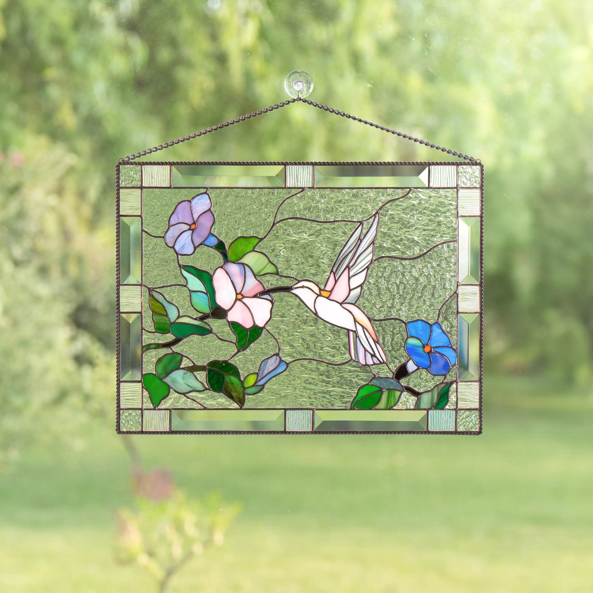 Stained glass panel with a hummingbird and flowers hanging against a blurred green background