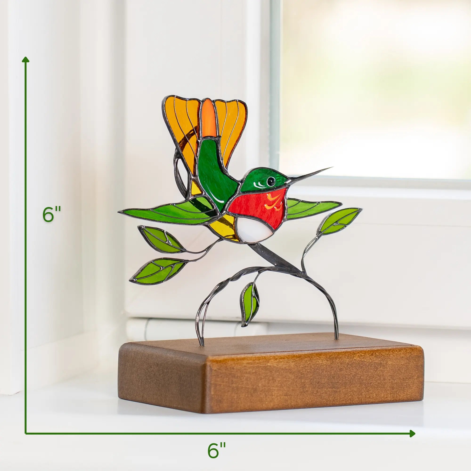 Stained glass hummingbird decor, size 6x6 inches, perched on branch with leaves and set on wooden base, shown on a windowsill with green accents.