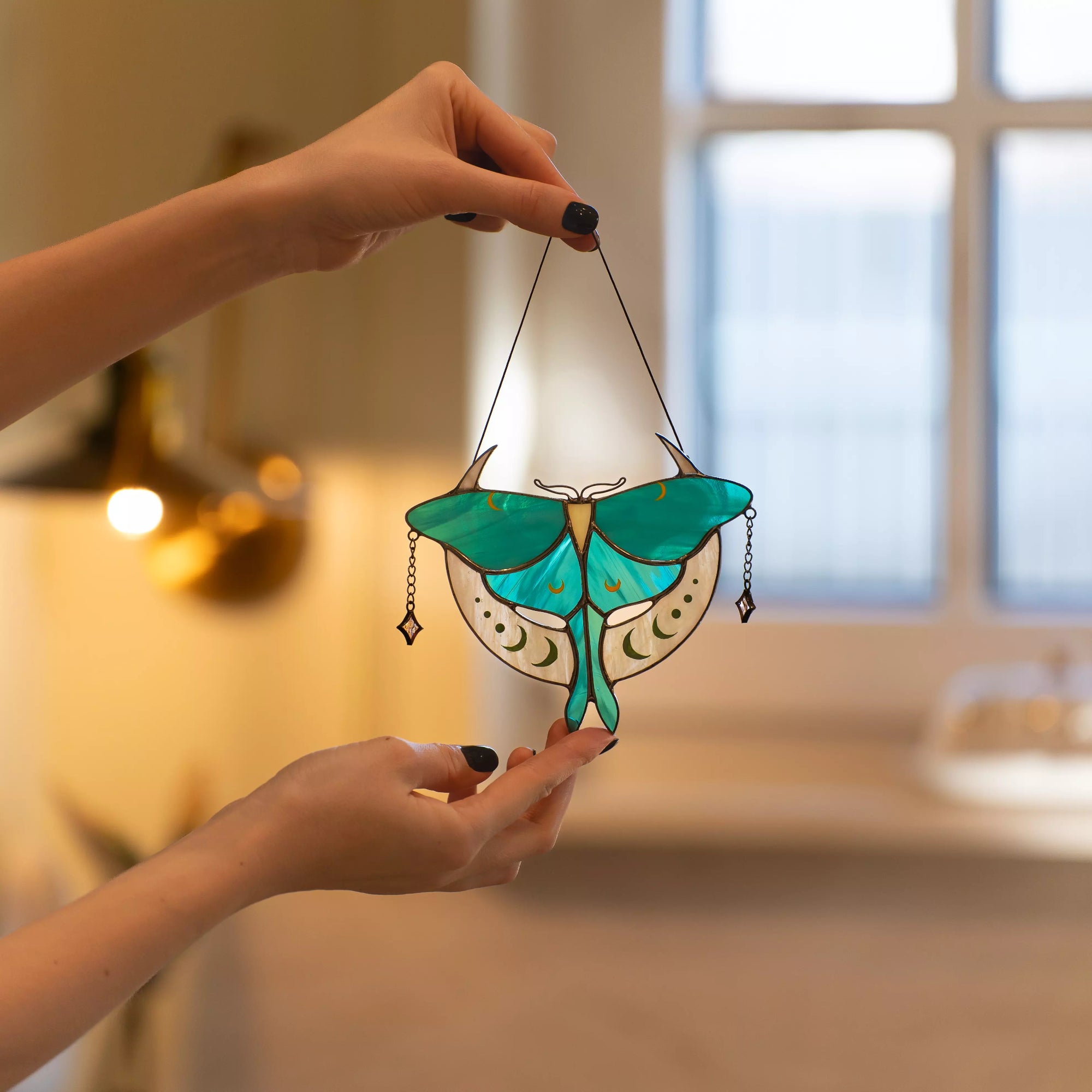 A person holding a Luna Moth stained glass suncatcher in a warm, cozy room near a window.