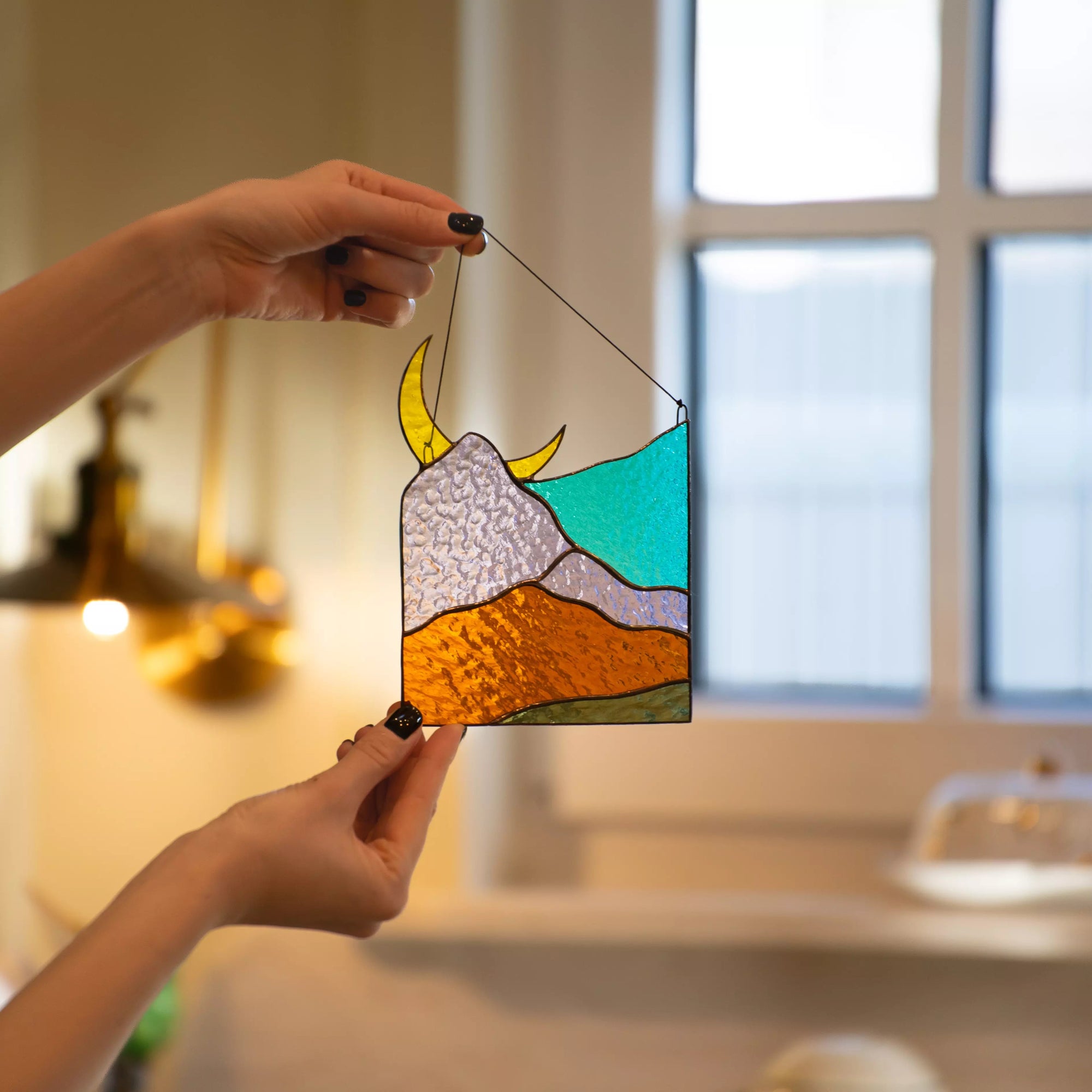 A person holding a mountain moon stained glass suncatcher in a warm, cozy room near a window.