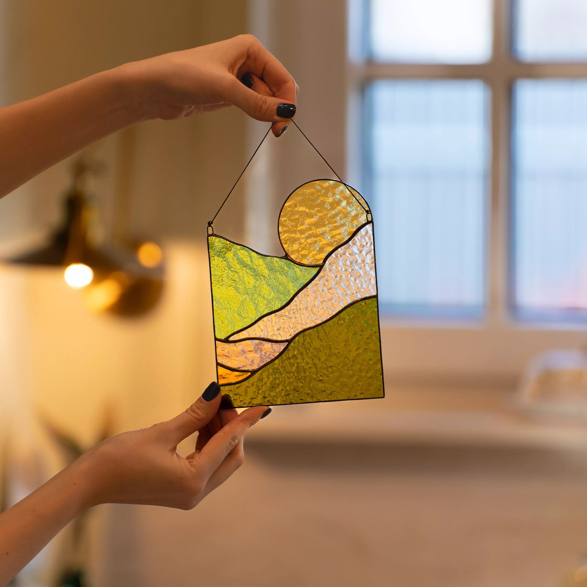 A hand holding a sun and mountain stained glass suncatcher in a cozy, sunlit interior setting.