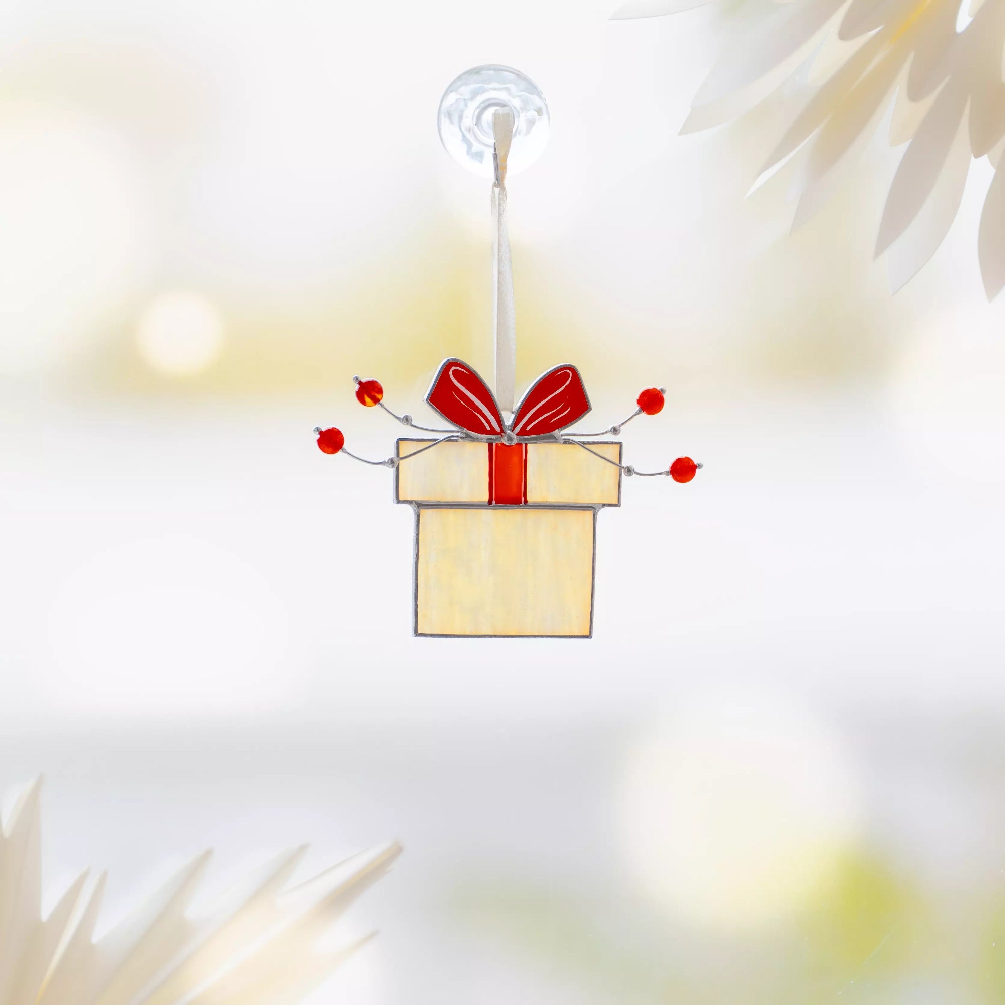 Iridescent stained glass Christmas gift ornament with red bow and red beads, hanging by a white ribbon on a window