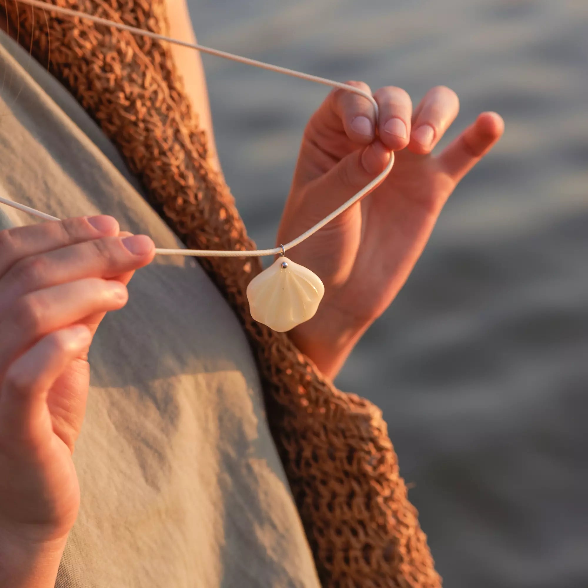 Person holding a necklace with a shell stained glass pendant by a body of water