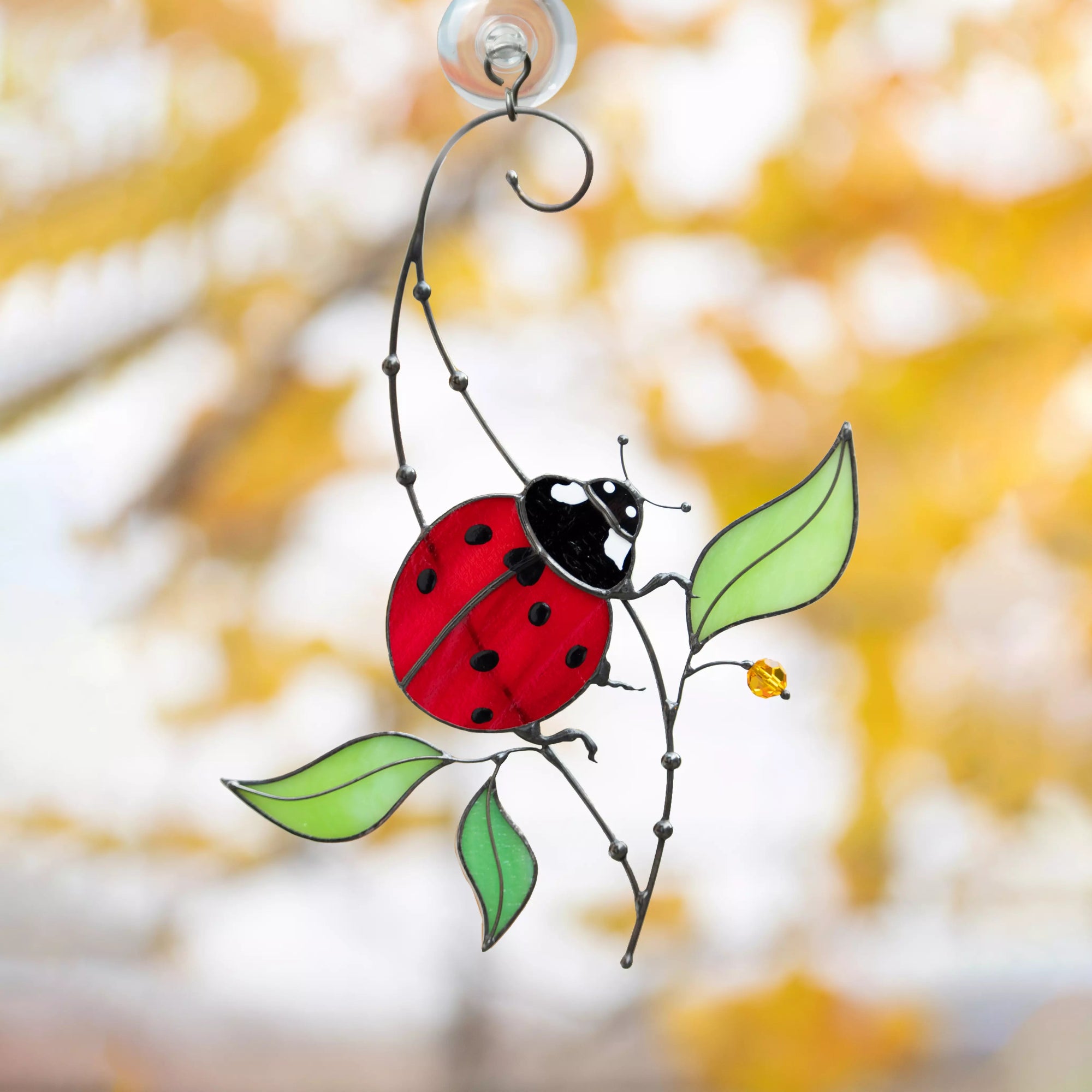 Close-up of a ladybug stained glass ornament with red wings, black spots, and green leaves hanging with a metal swirl design.