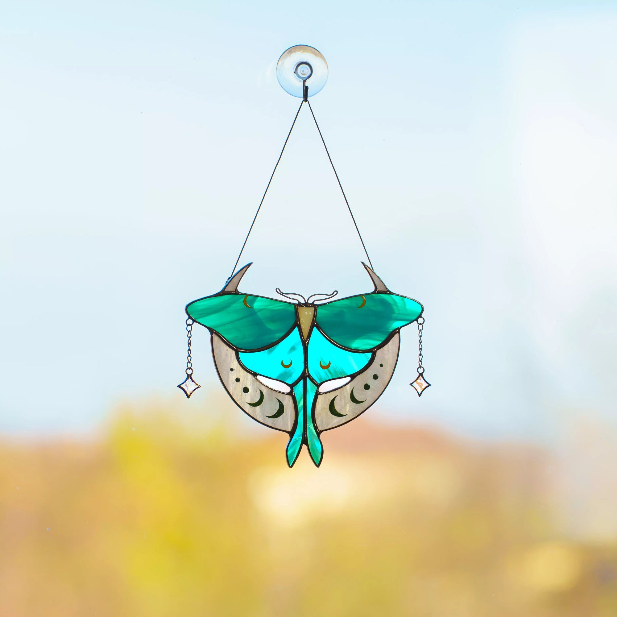 A hand holding a Luna Moth stained glass suncatcher that casts a beautiful teal and grey shadow.
