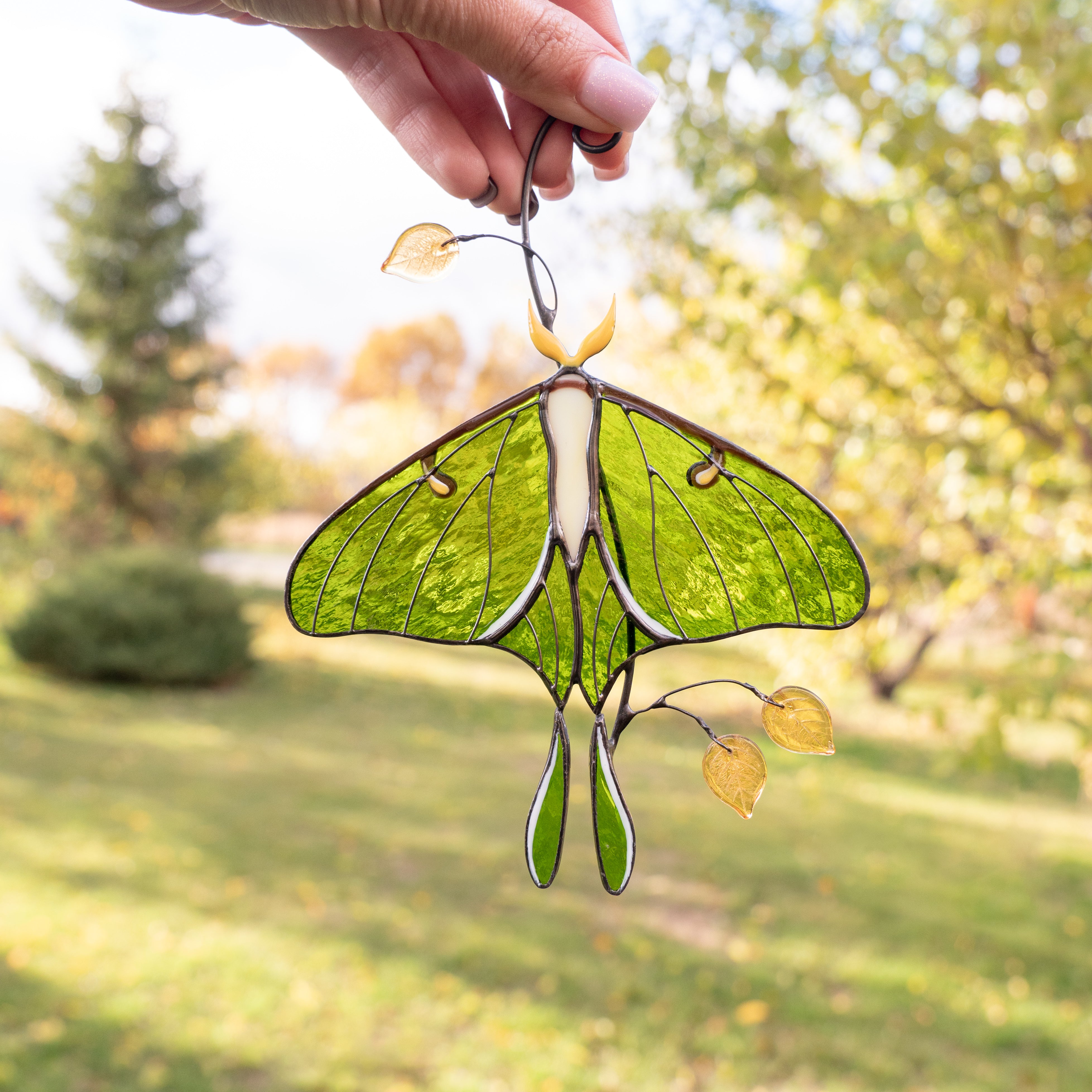 Green stained glass luna moth hanging on a window with blurred garden background.