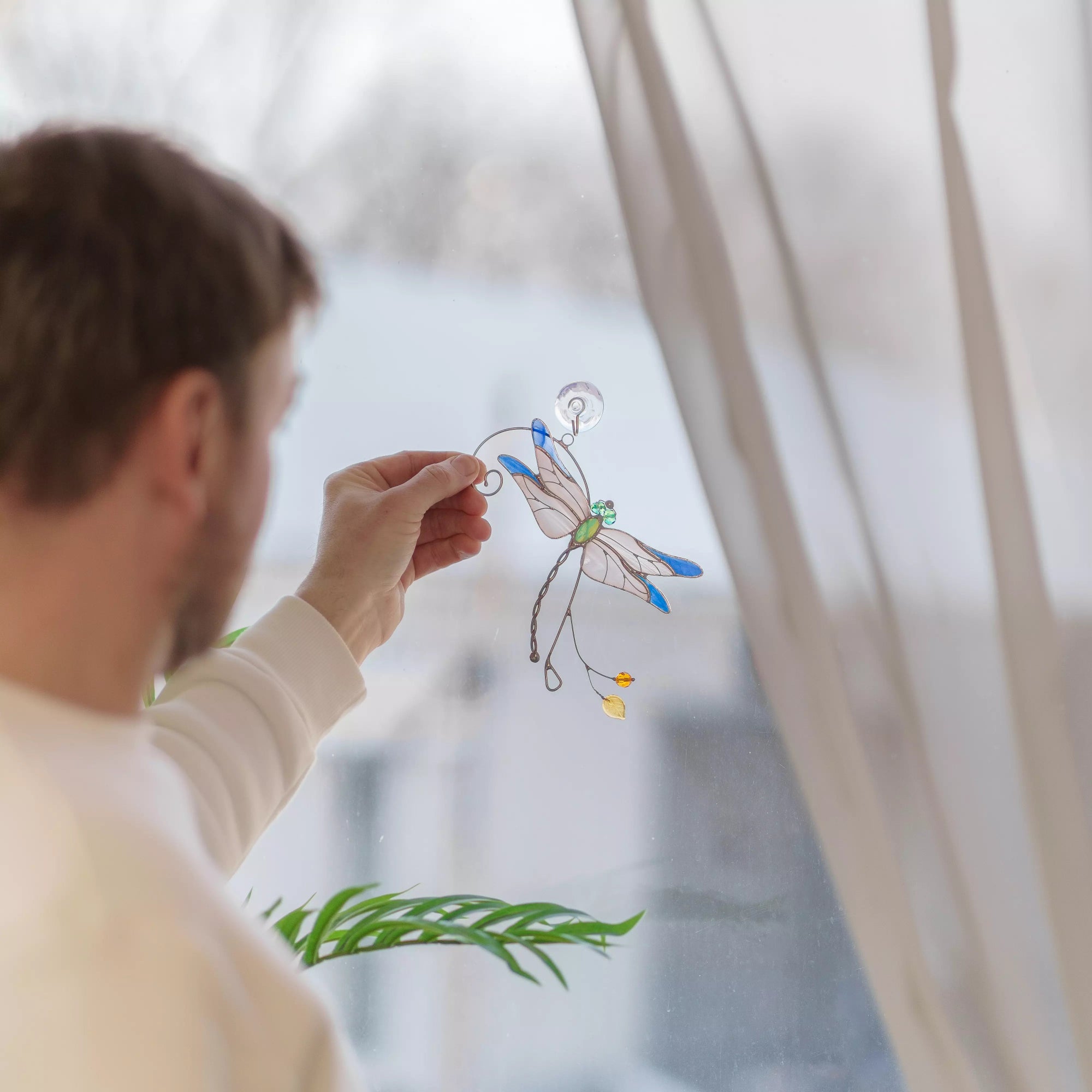 Person holding a stained glass dragonfly suncatcher near a window indoors