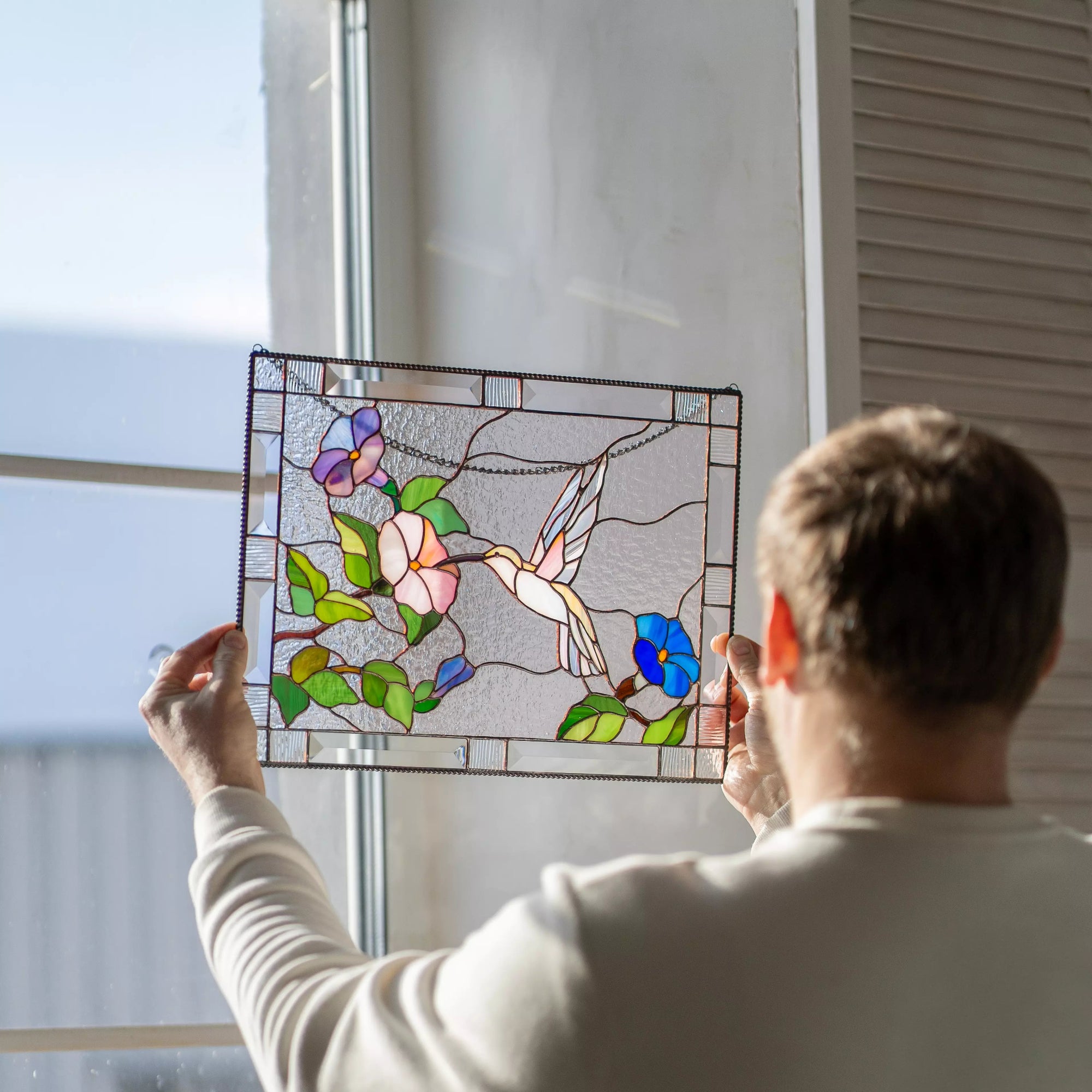 Person holding a stained glass panel with floral and bird design by a window