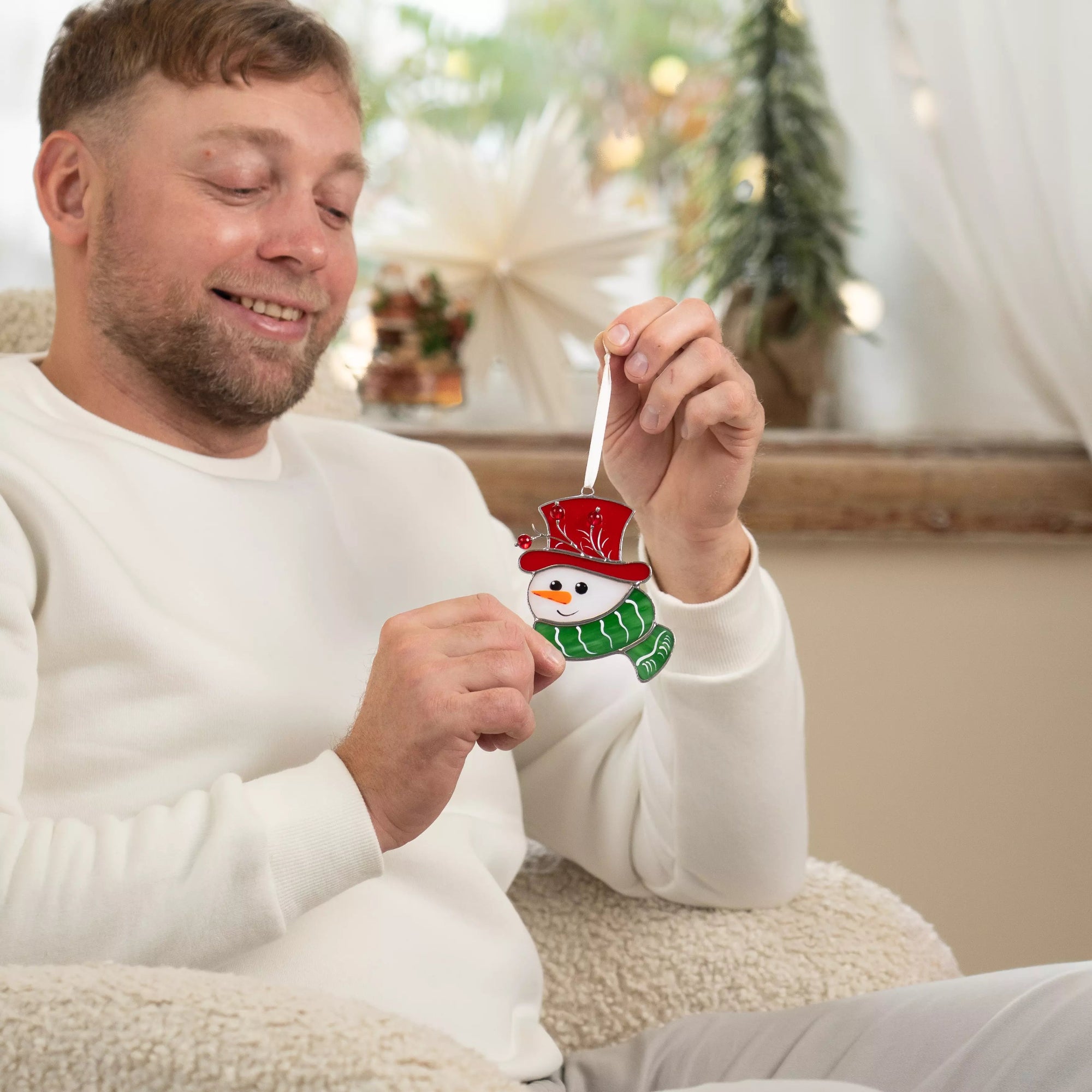 Man in white sweater smiling while holding a stained glass snowman ornament in cozy holiday room.