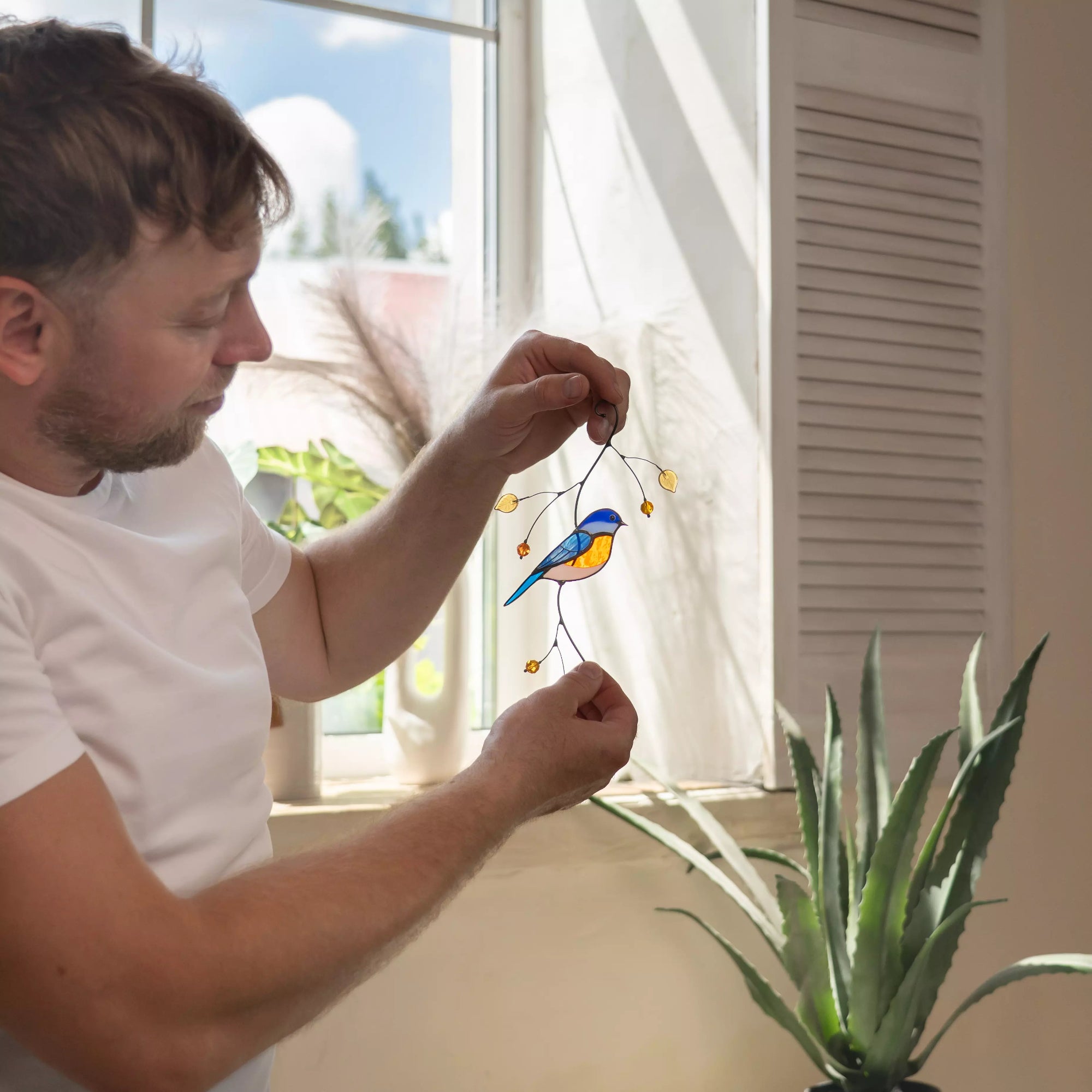 Man holding stained glass bluebird suncatcher with golden leaves, indoors with a window and plant in the background