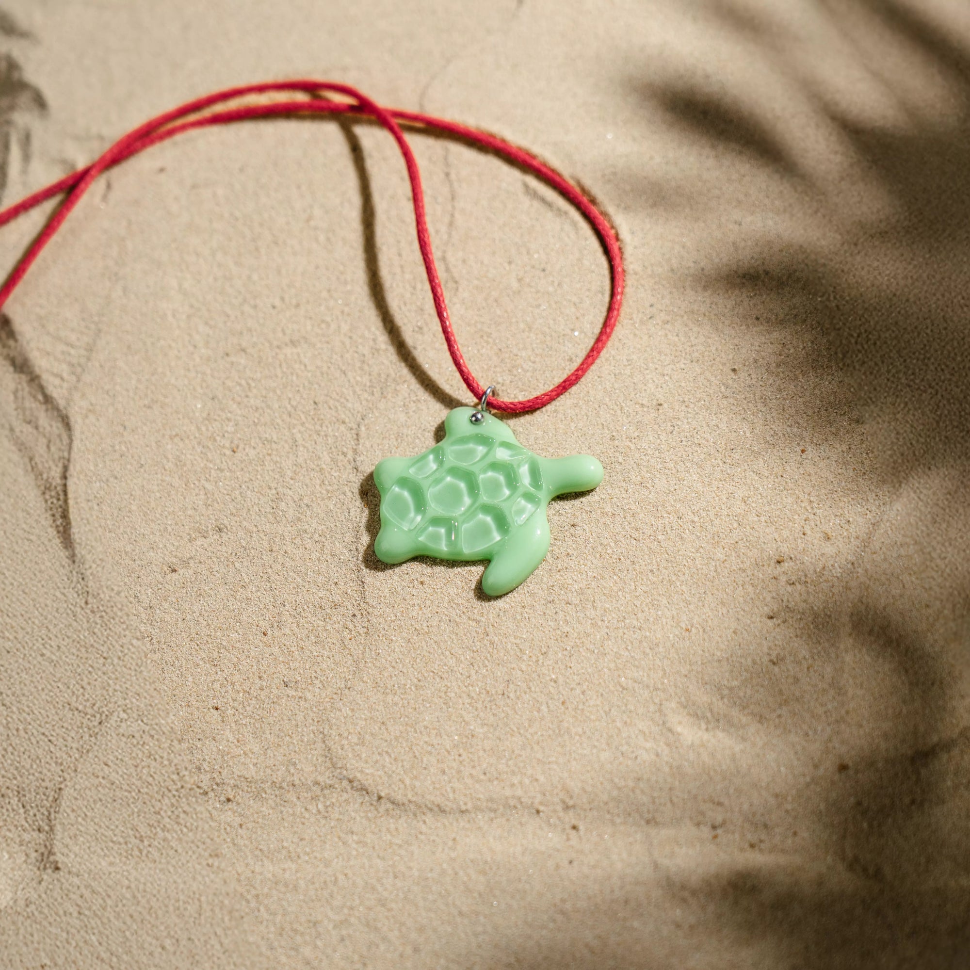 Mint green stained glass turtle necklace on a red string necklace lying on sand