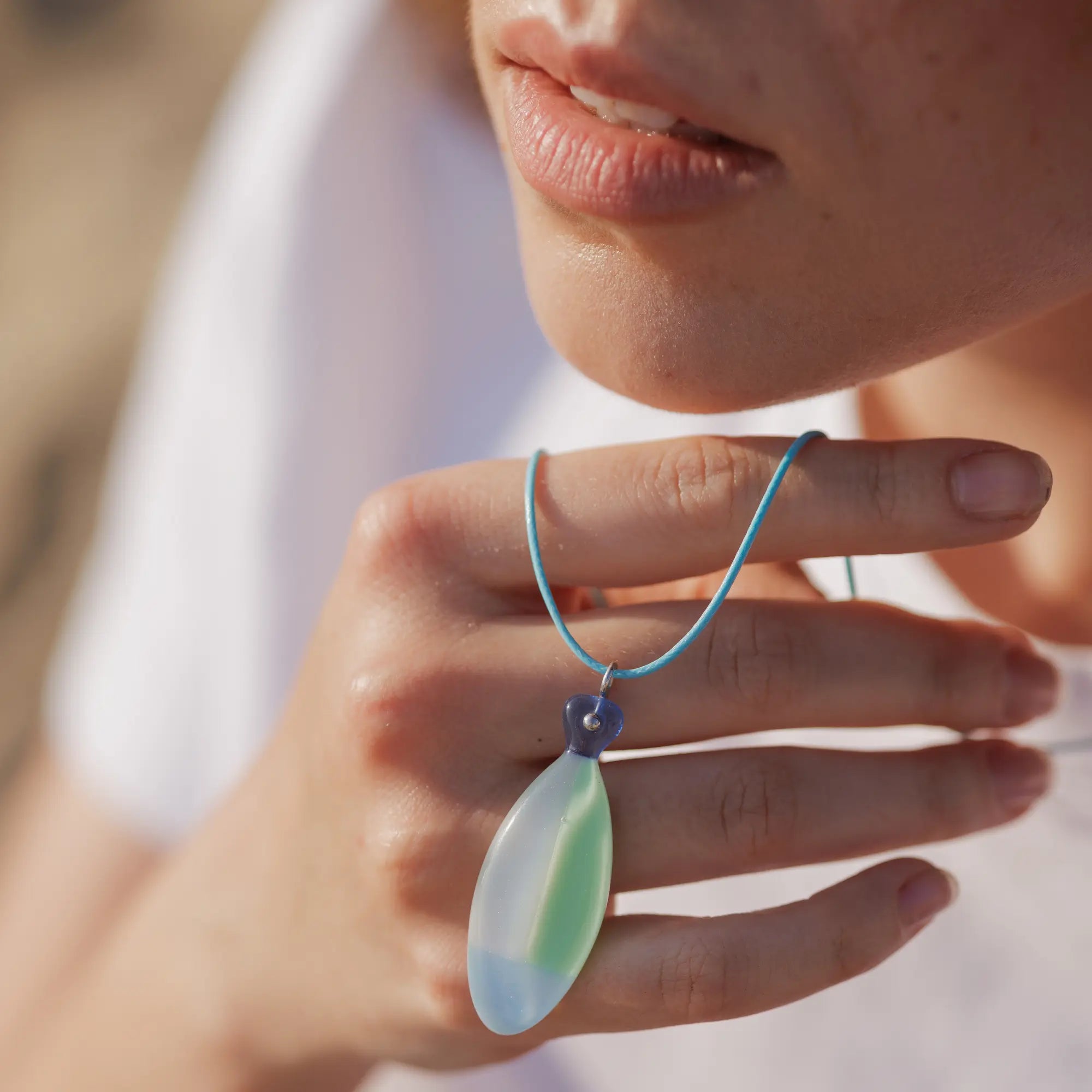 Close-up of hand holding green and blue glass fish necklace with turquoise cord, worn by a woman outdoors.