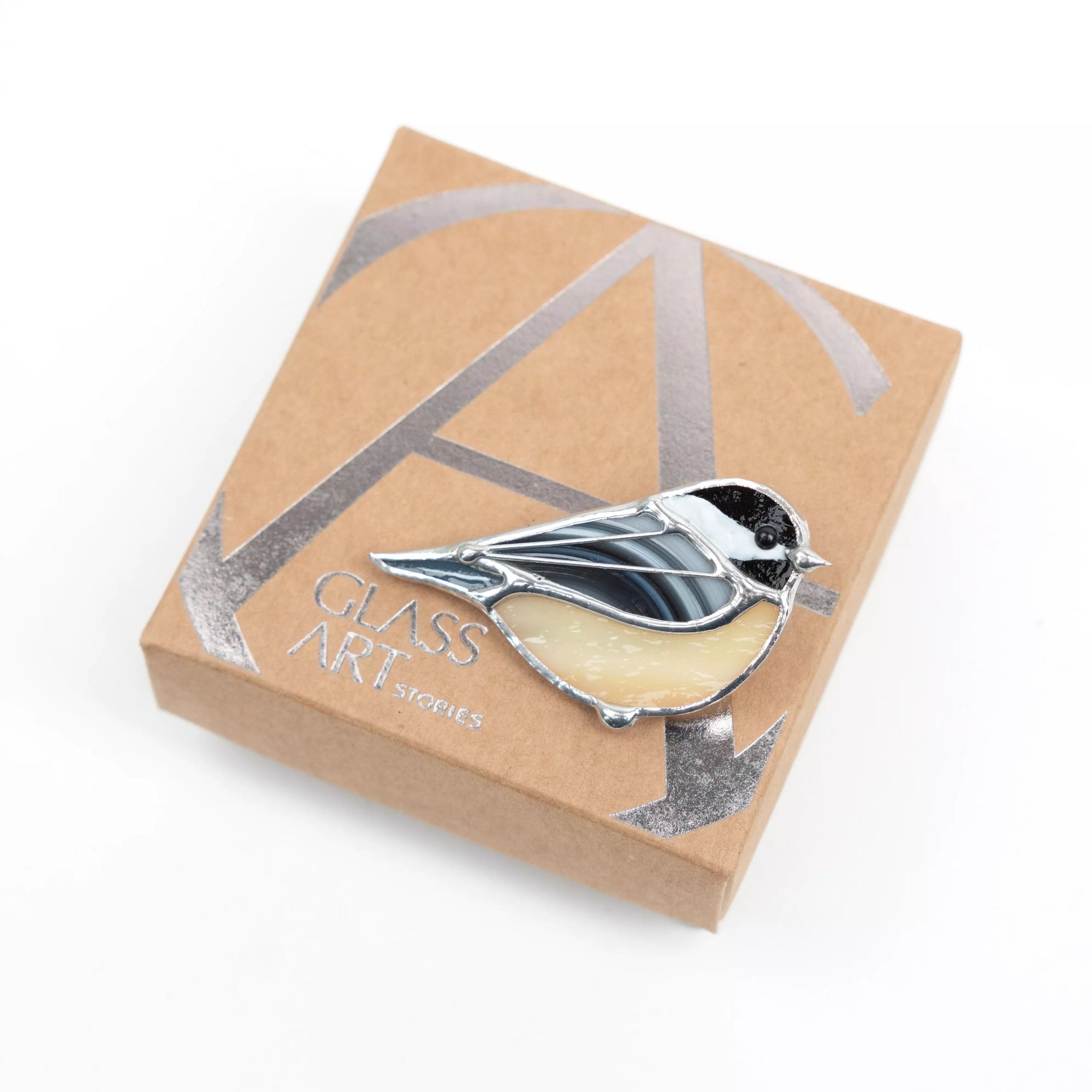 Cream and black bird brooch made of stained glass placed diagonally on branded kraft gift box.