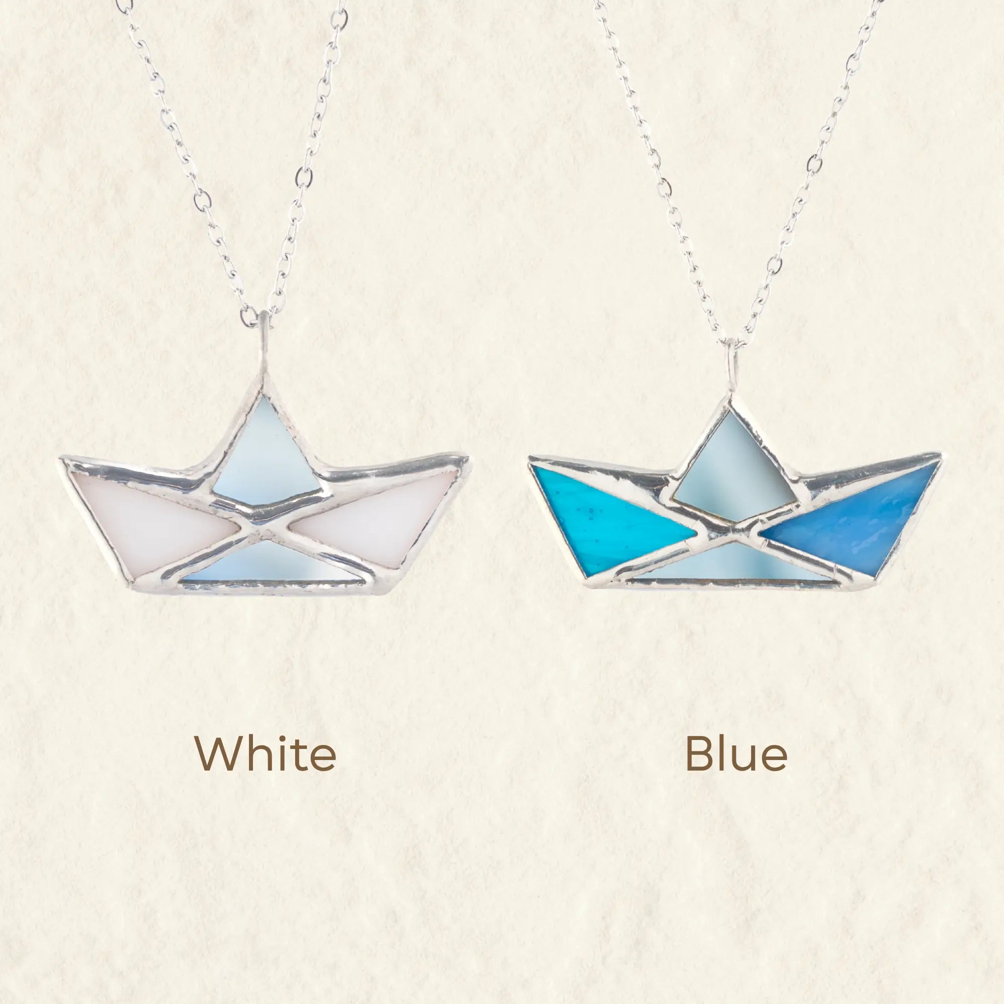 Two paper boat stained glass pendant necklaces on silver chains, labeled White and Blue, displayed side by side on a textured cream background.