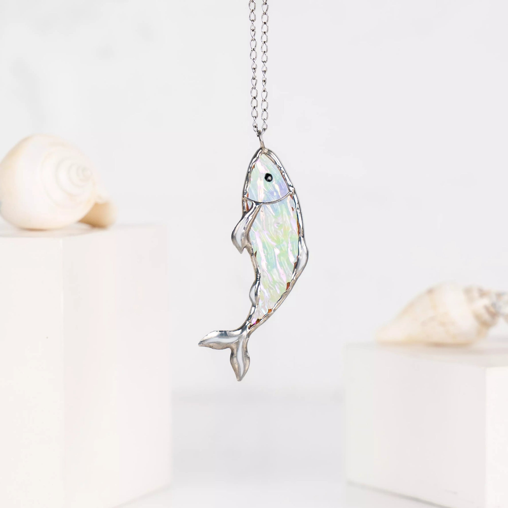 Clear stained glass fish necklace in light tones, shown with soft focus seashells in the background