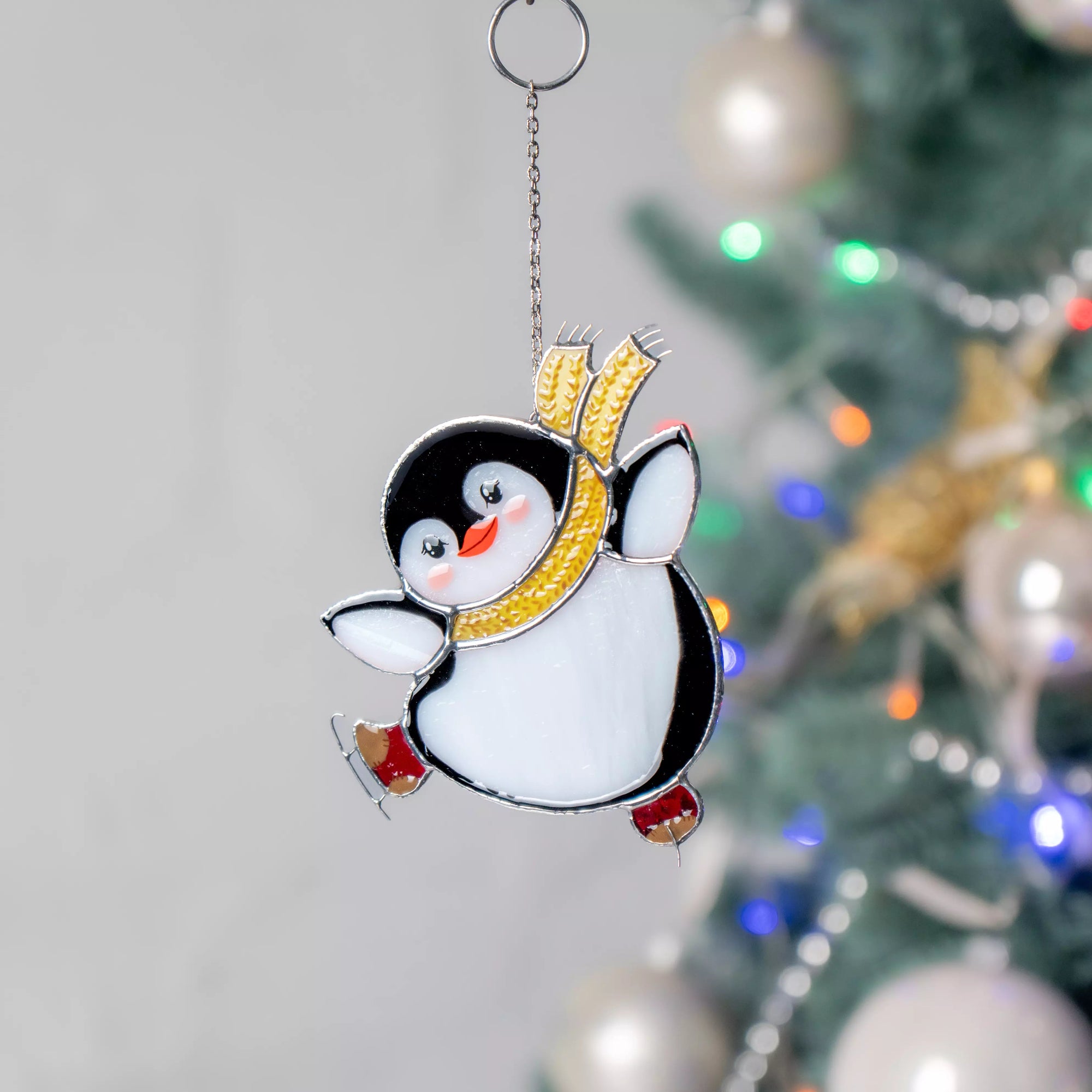 Penguin stained glass suncatcher with a yellow scarf hanging in front of a decorated Christmas tree.