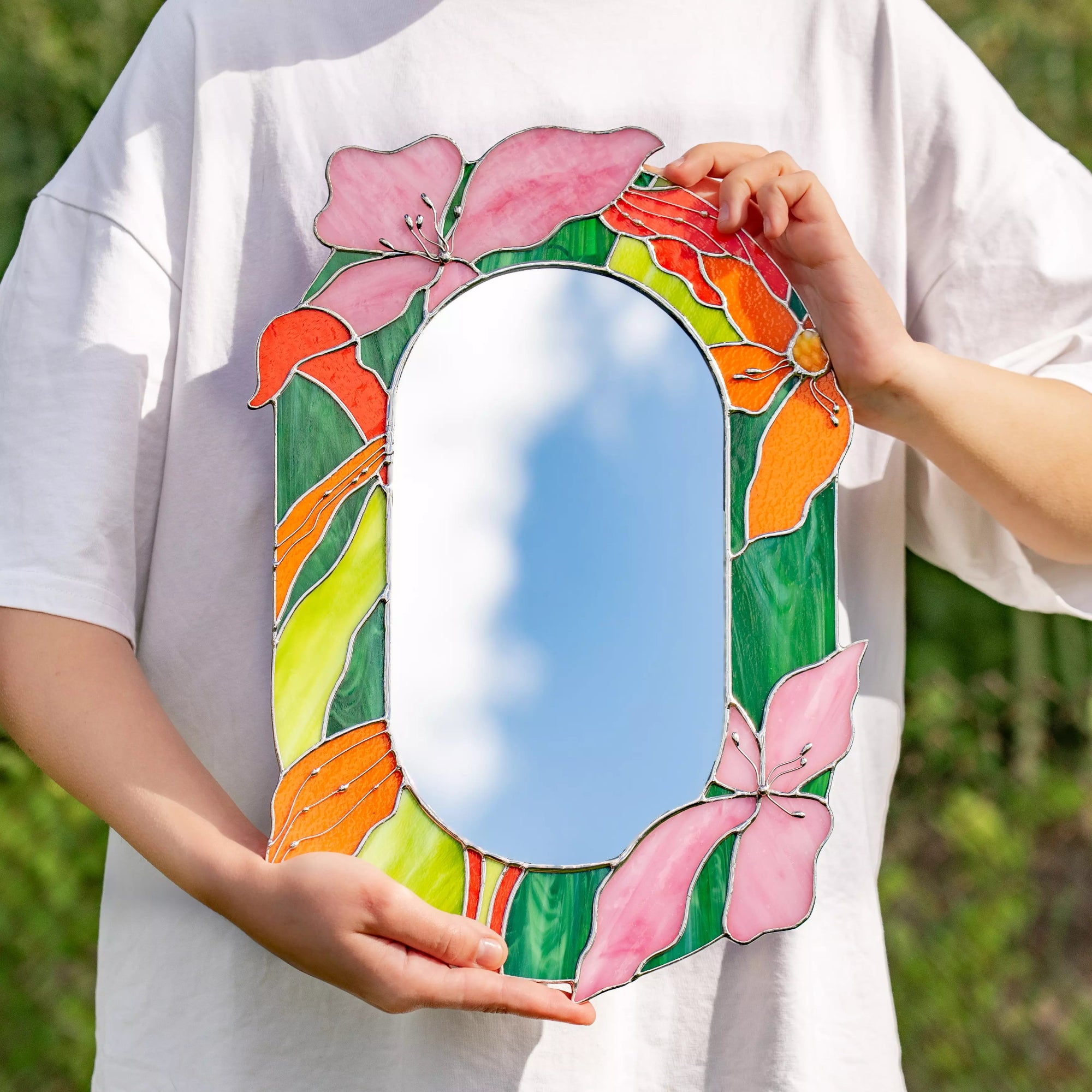 Person holding a colorful stained glass floral mirror against a white shirt outdoors.