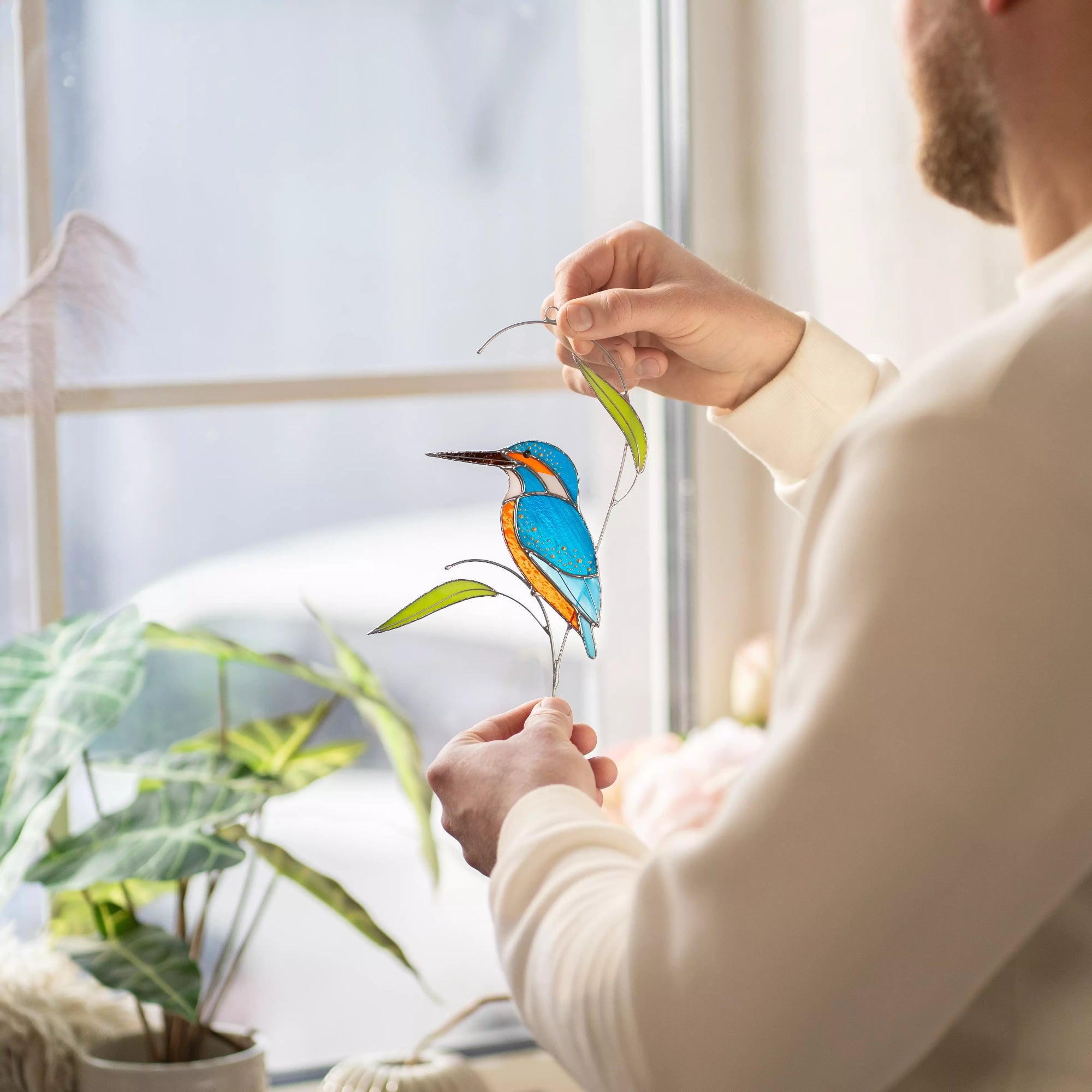 Man holding a colorful stained glass kingfisher suncatcher in front of a window