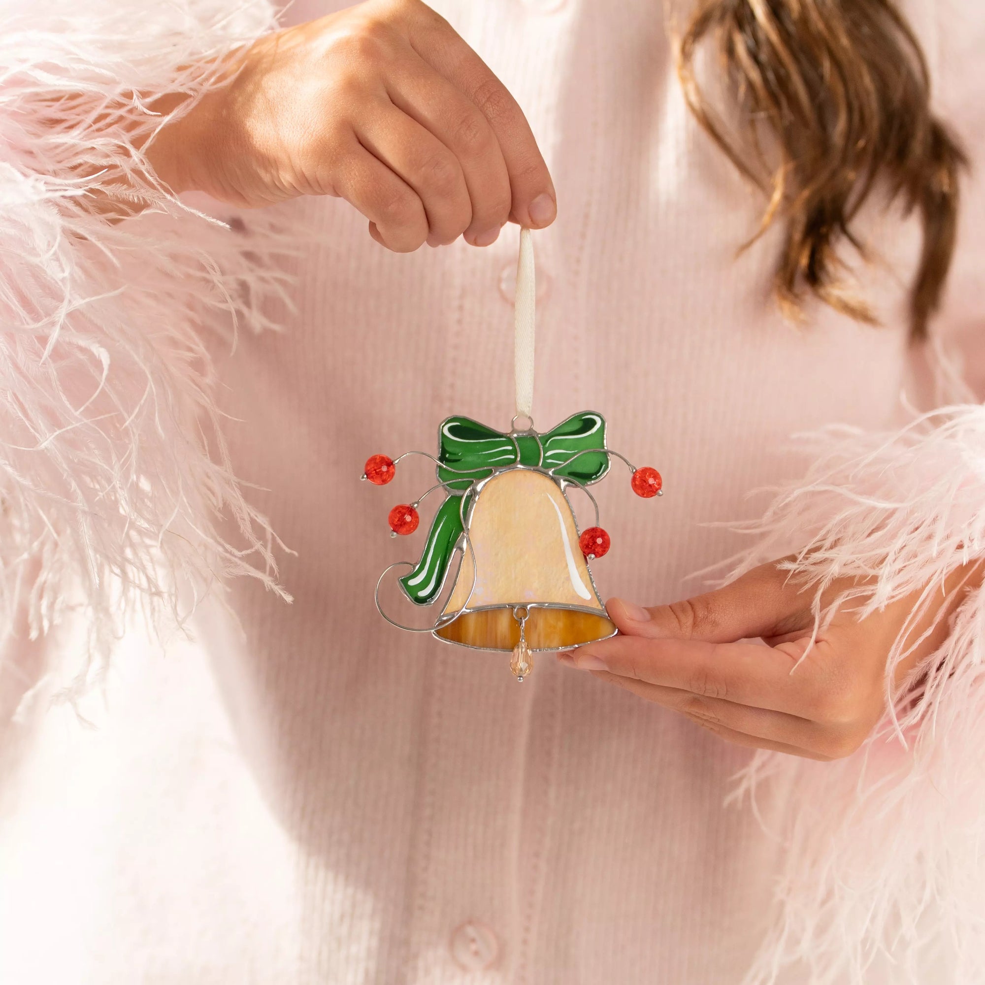 Person holding a decorative bell stained glass Christmas ornament with a green bow and red beads.