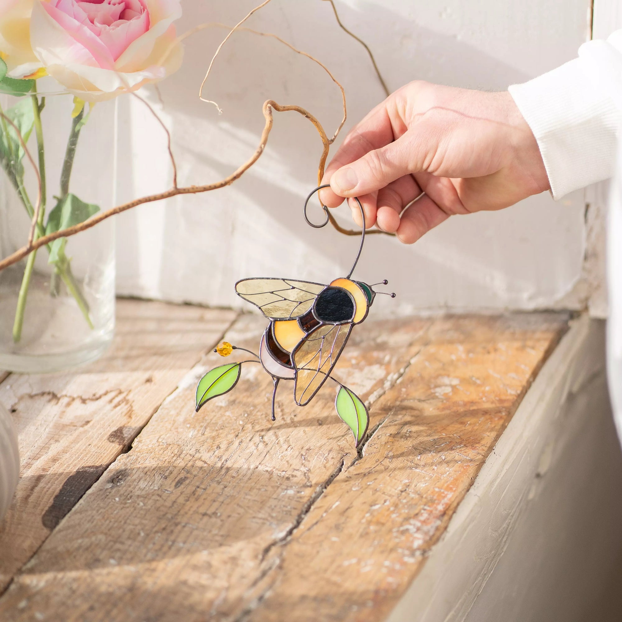 Hand holding a stained glass suncatcher bumble bee on a wooden surface with flowers in the background