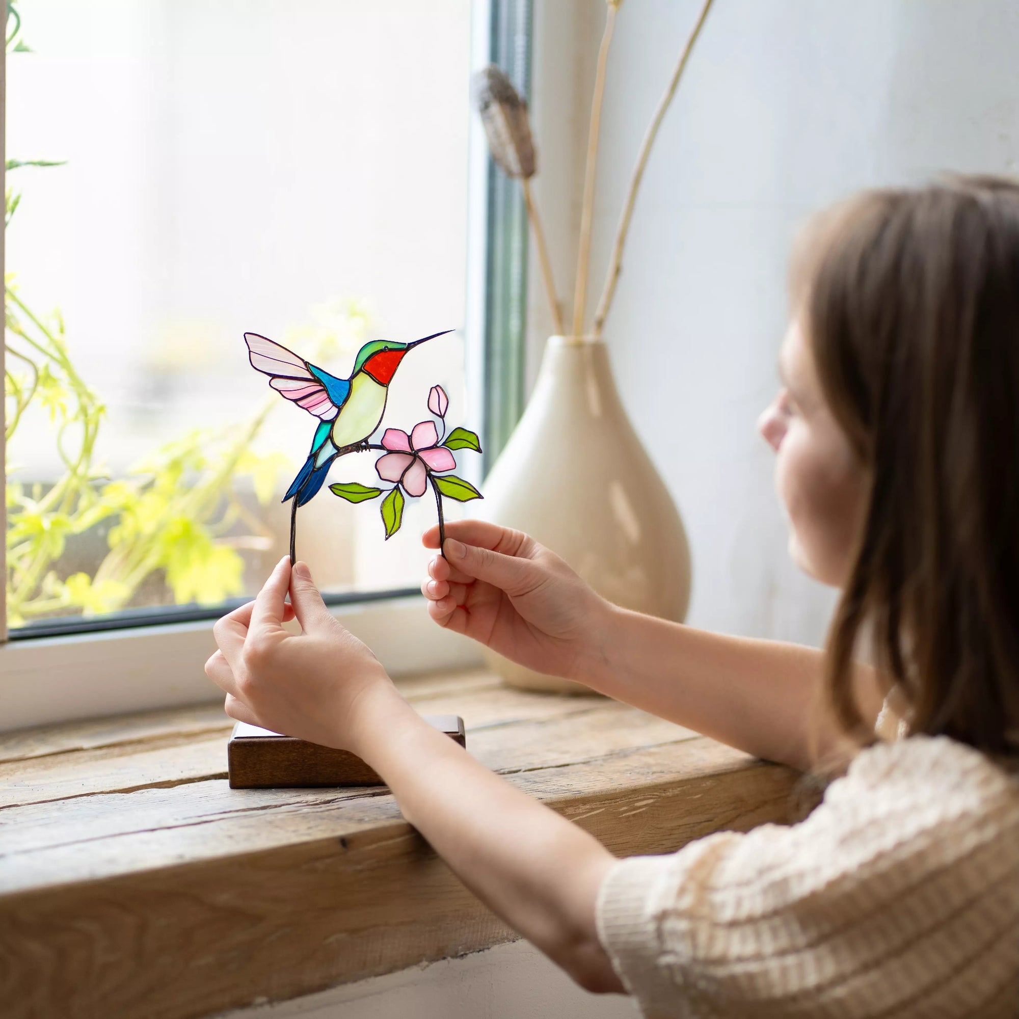 Women holding a stained glass hummingbird and flower suncatcher by a window