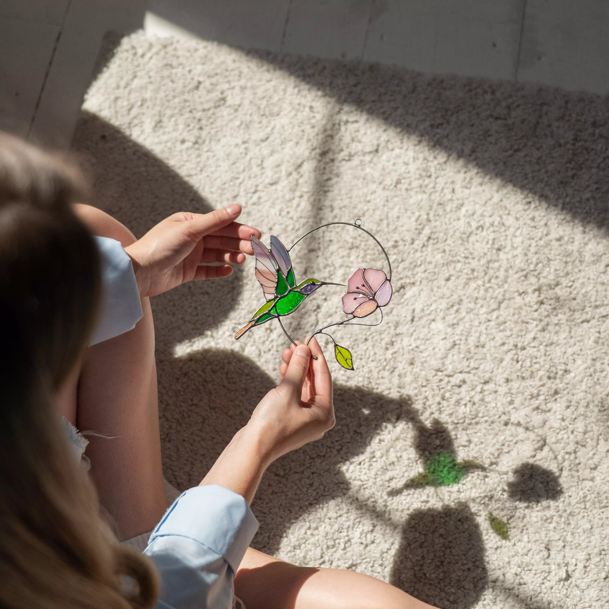Person holding a stained glass hummingbird and lotus suncatcher with soft shadow on the floor
