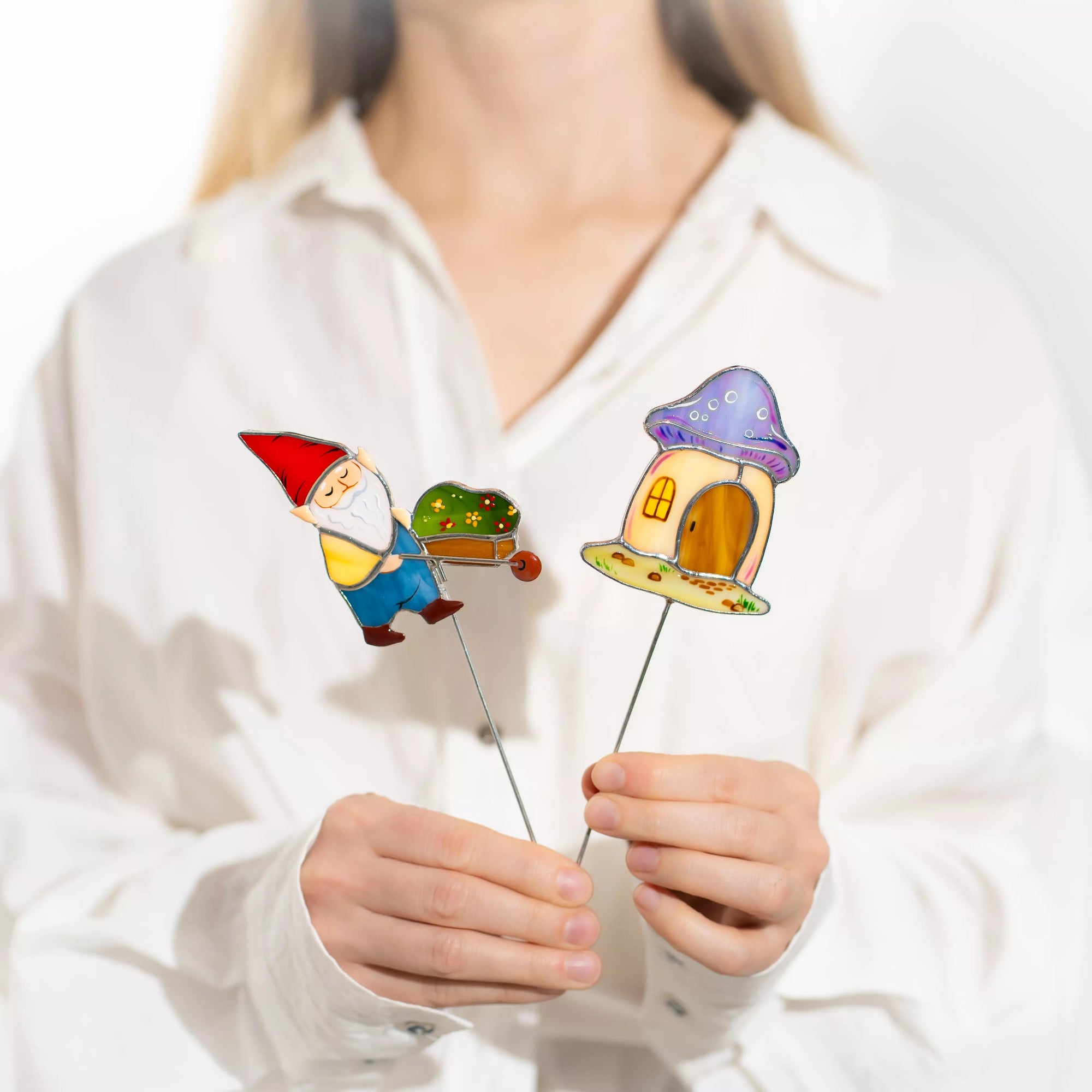 Hands holding two colorful plant stakes with a gnome and mushroom house design.