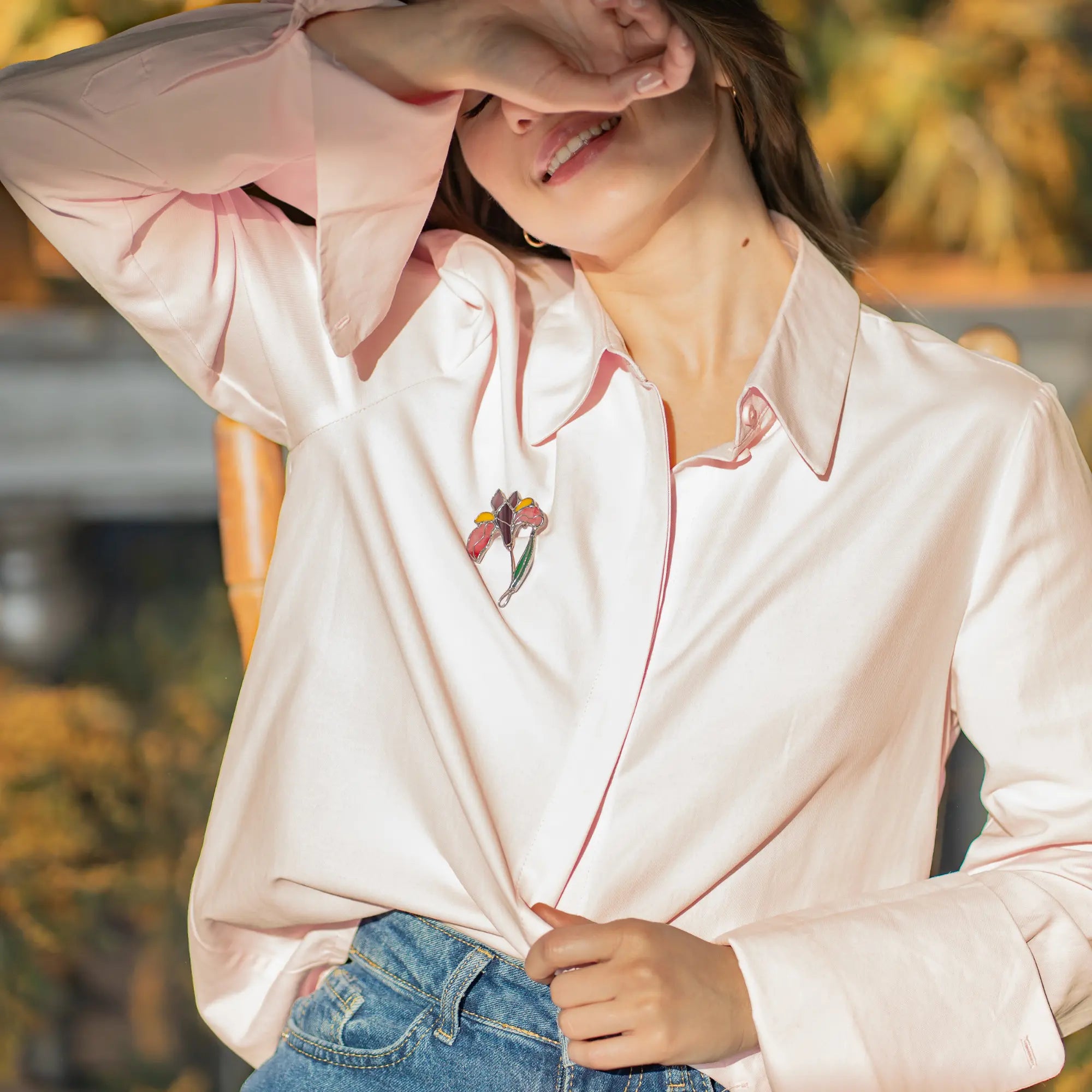 Woman wearing a light pink blouse with floraliris brooch and blue jeans outdoors.