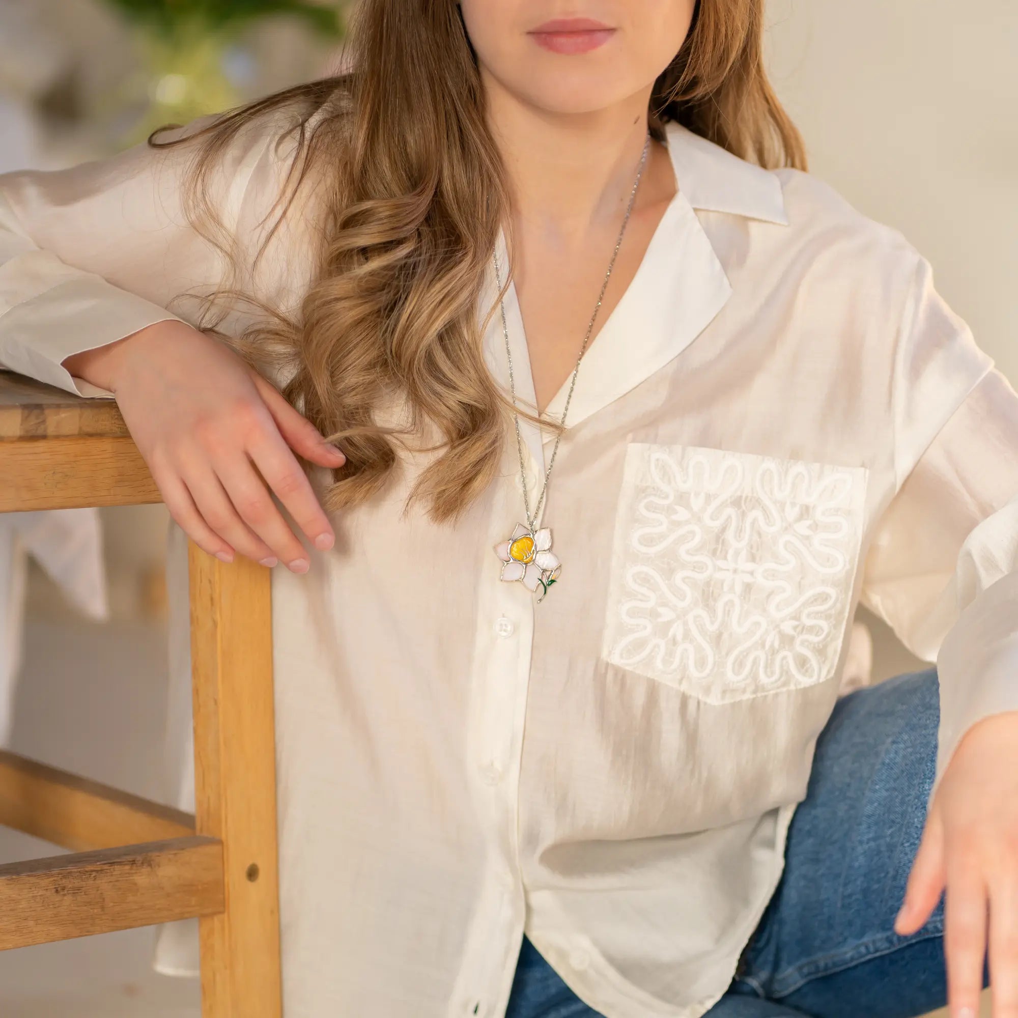 Woman wearing a white blouse with floral stained glass brooch sitting on a wooden chair.