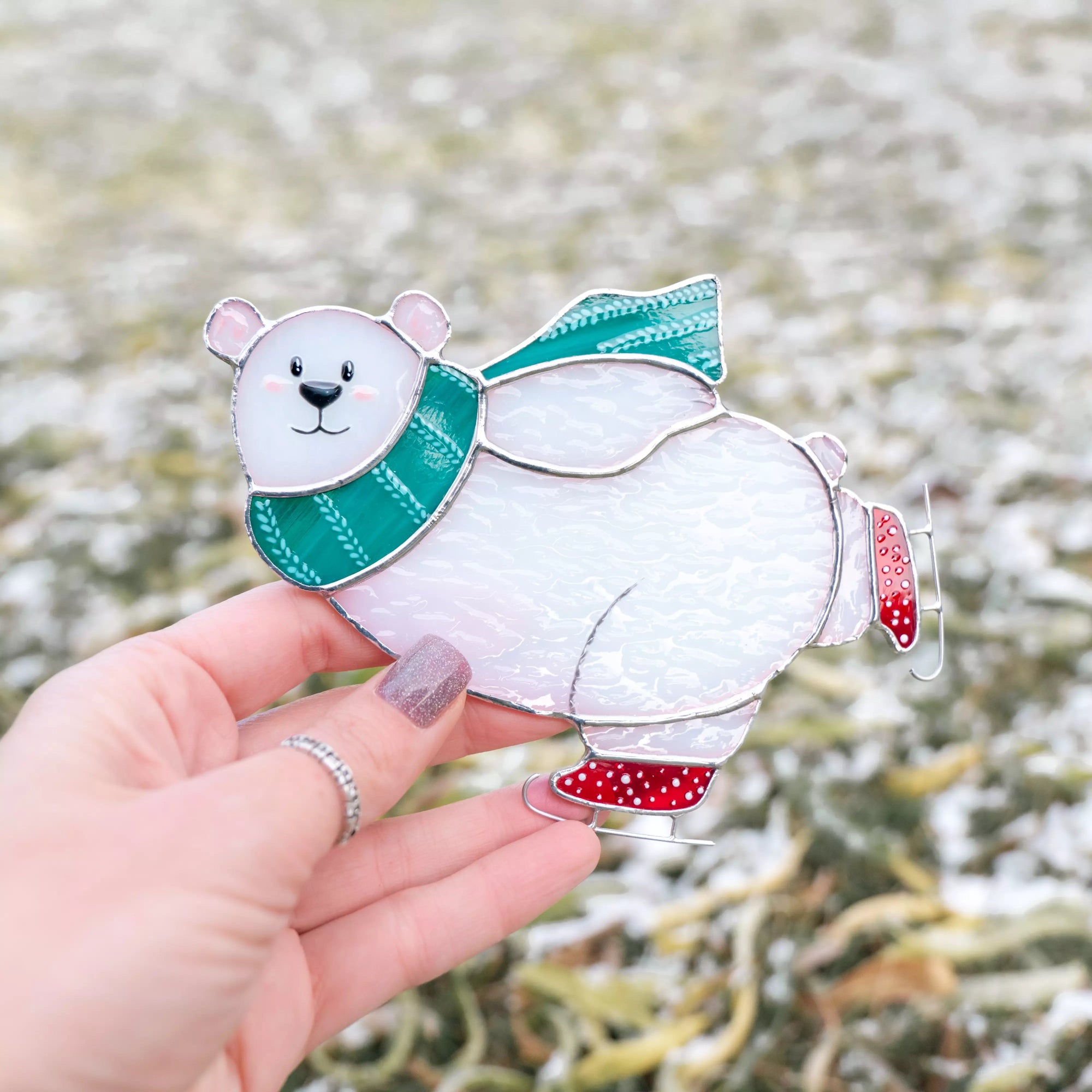 Polar bear stained glass ornament with a green scarf and red shoes held by a hand against a blurred snowy background.