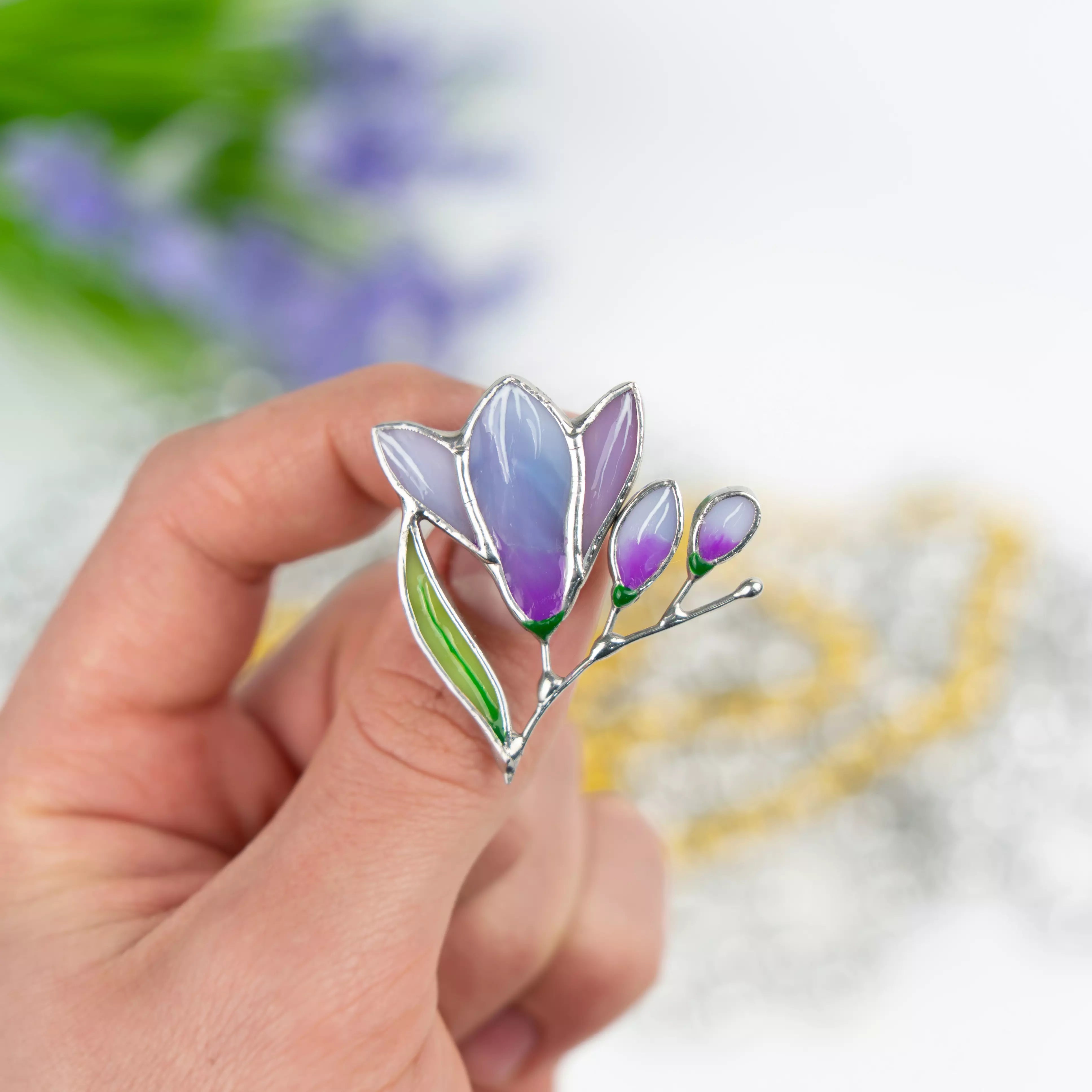 Hand holding a stained glass brooch shaped like a purple freesia flower with green leaf