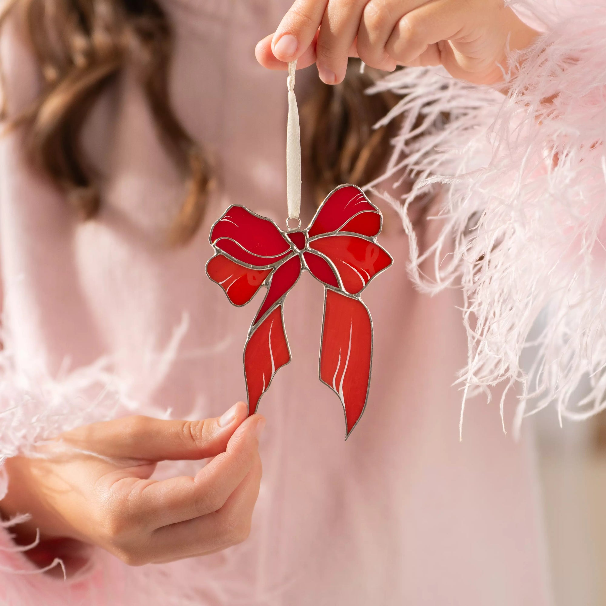 Red bow stained glass ornament held by a person wearing a pink feathered garment