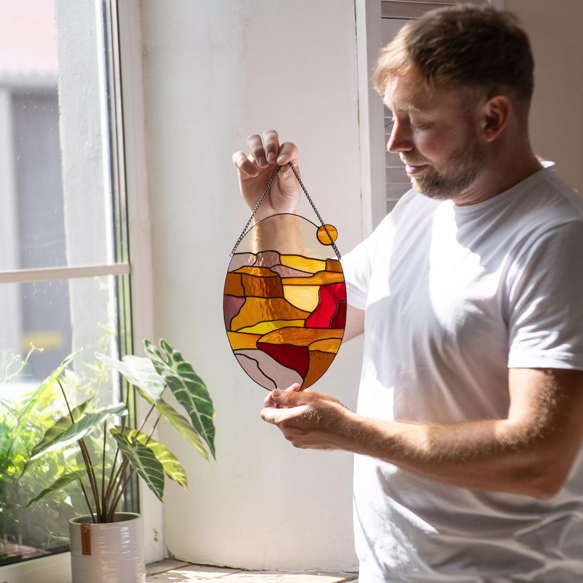 Man holding stained glass landscape panel near bright window with green plant nearby.