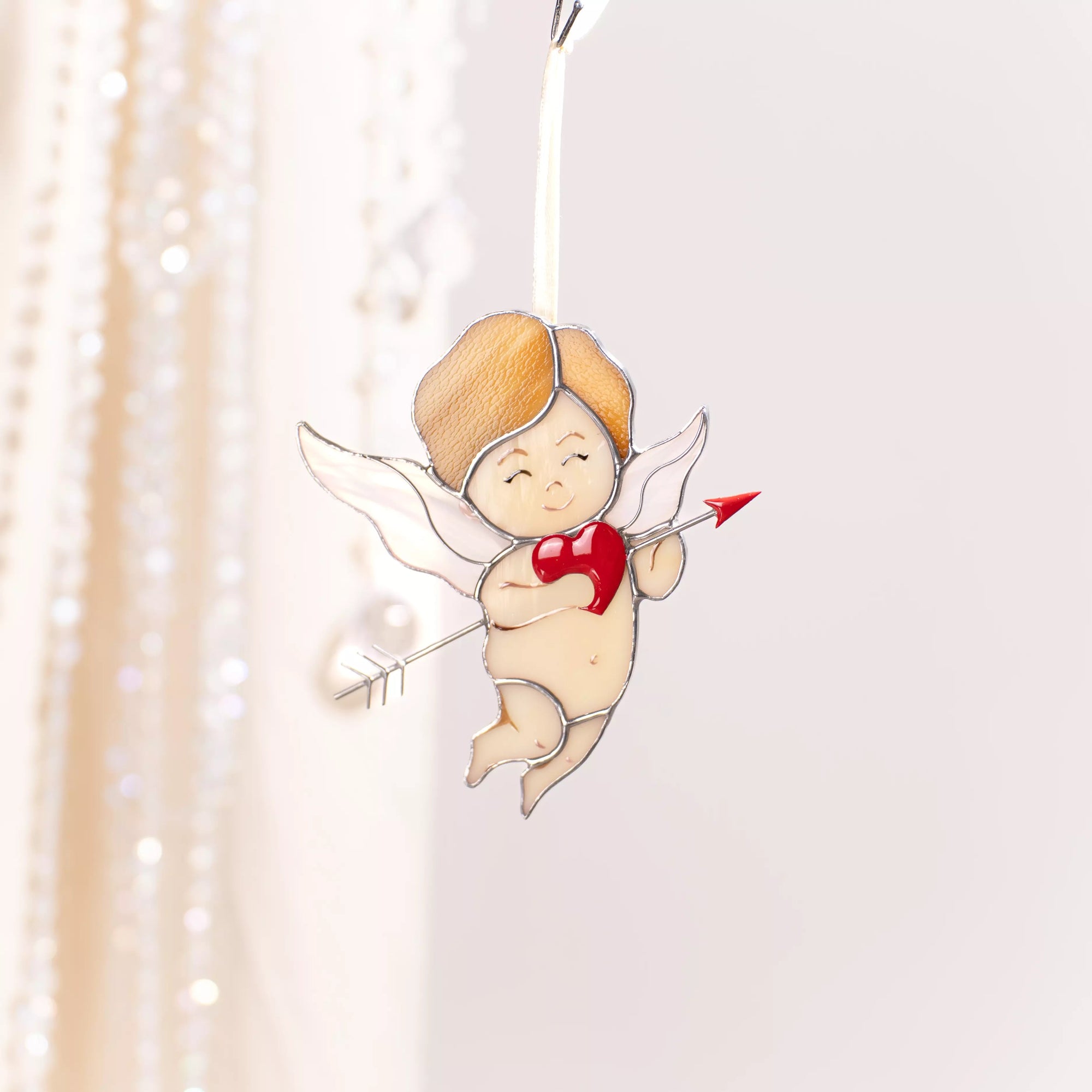 Stained glass cupid suncatcher hanging on a ribbon in a bright interior, glowing softly in natural light.