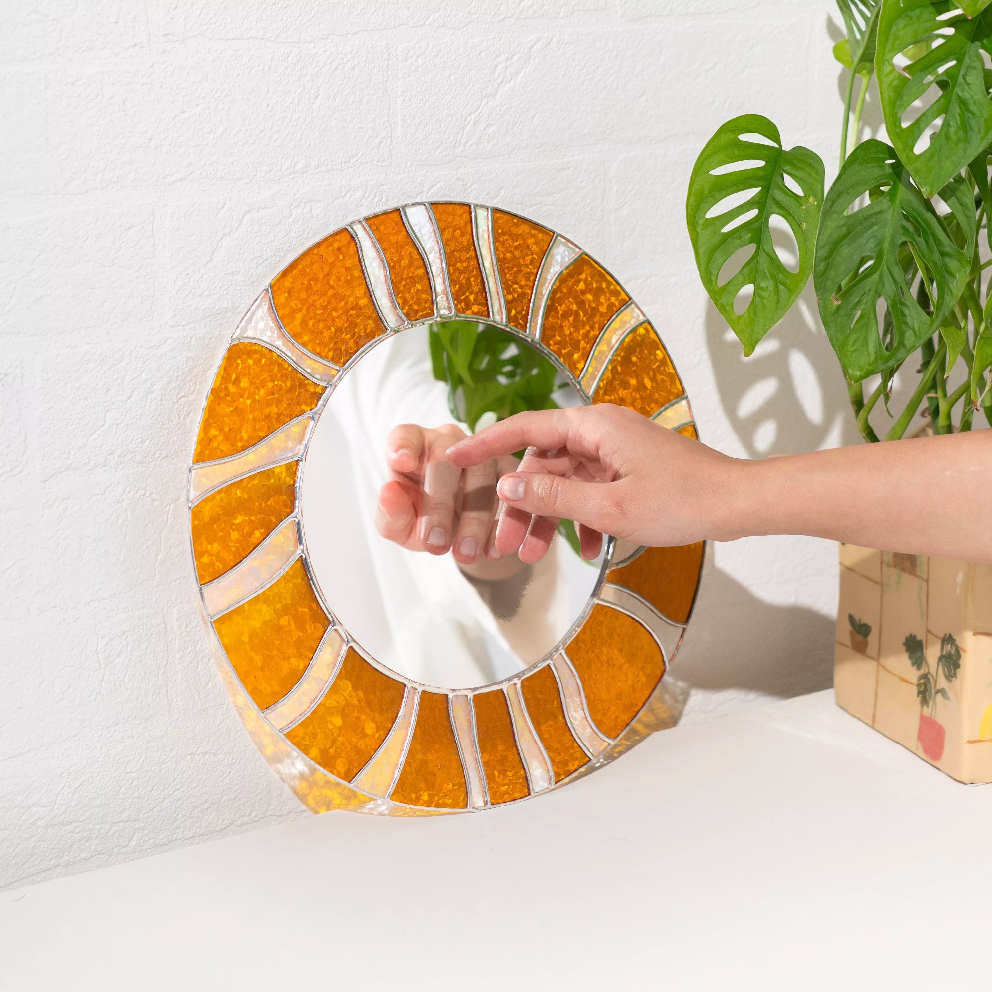 Round mirror with orange and brown frame on a white surface with a plant in the background