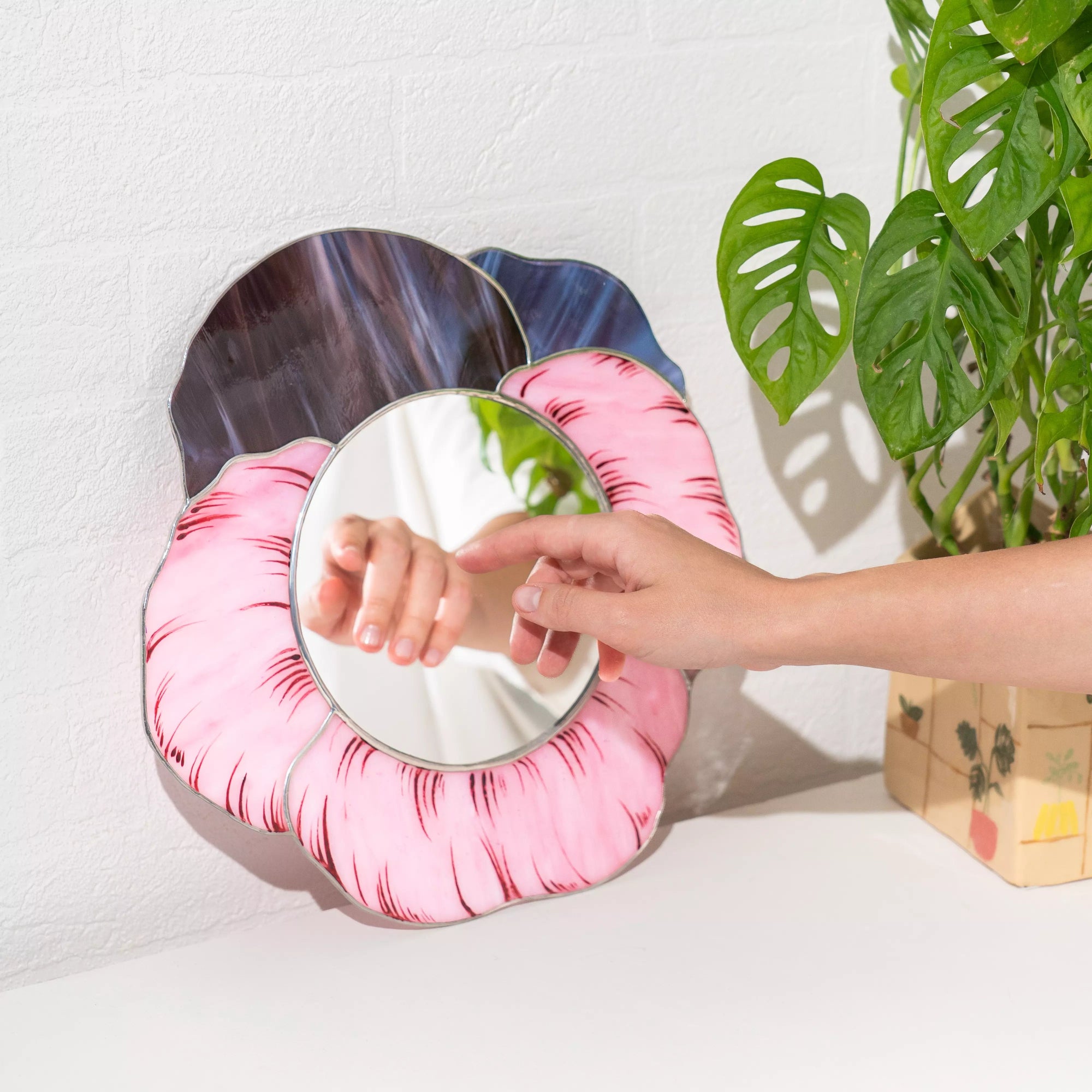 Round stained glass mirror with pink and blue floral frame held by a hand against a white wall with a plant.