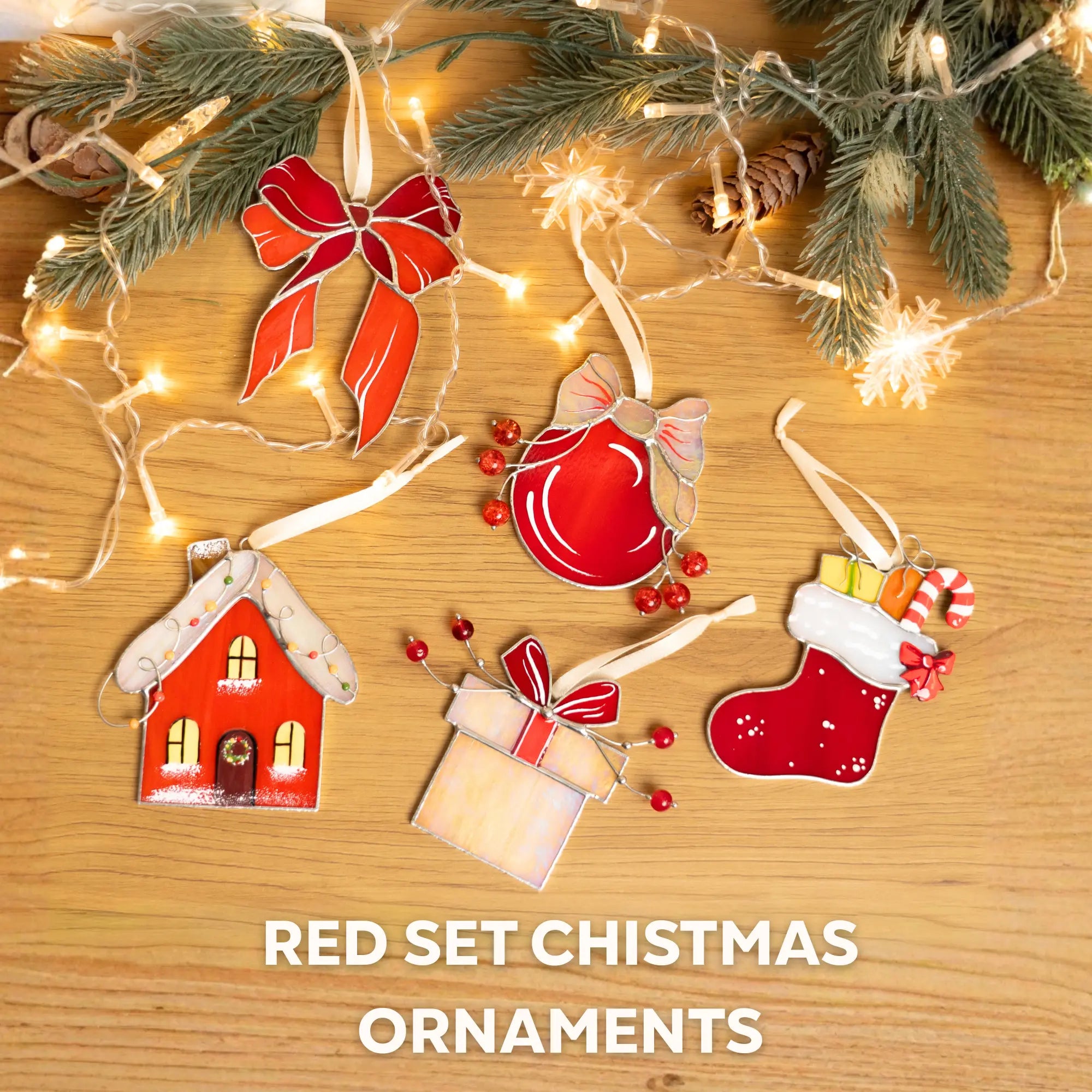 Set of red stained glass Christmas ornaments featuring: gingerbread house, red ball, red bow, stocking and iridescent gif box -on a wooden surface with lights and greenery.