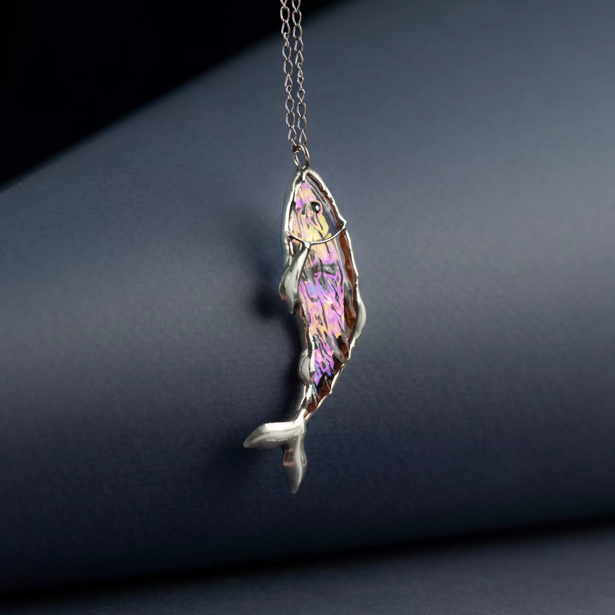 Vibrant irisated stained glass fish pendant with bright tones hanging in front of dark grey paper