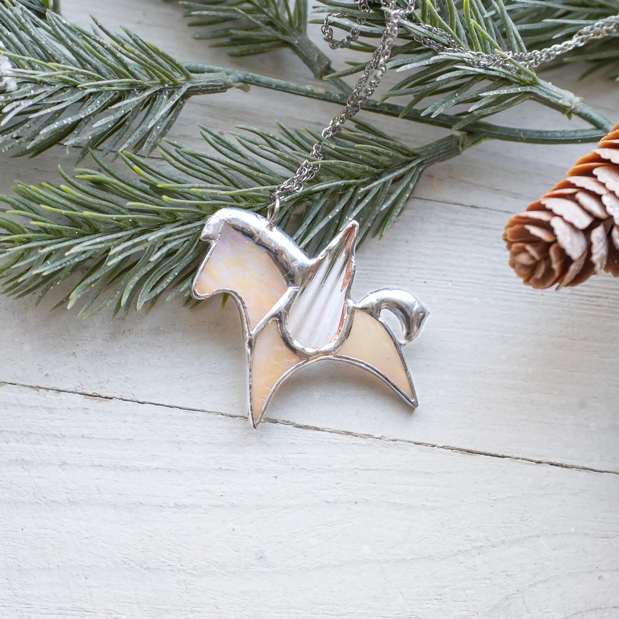Silver stained glass pegasus necklace on a white wooden surface with greenery and pine cones.