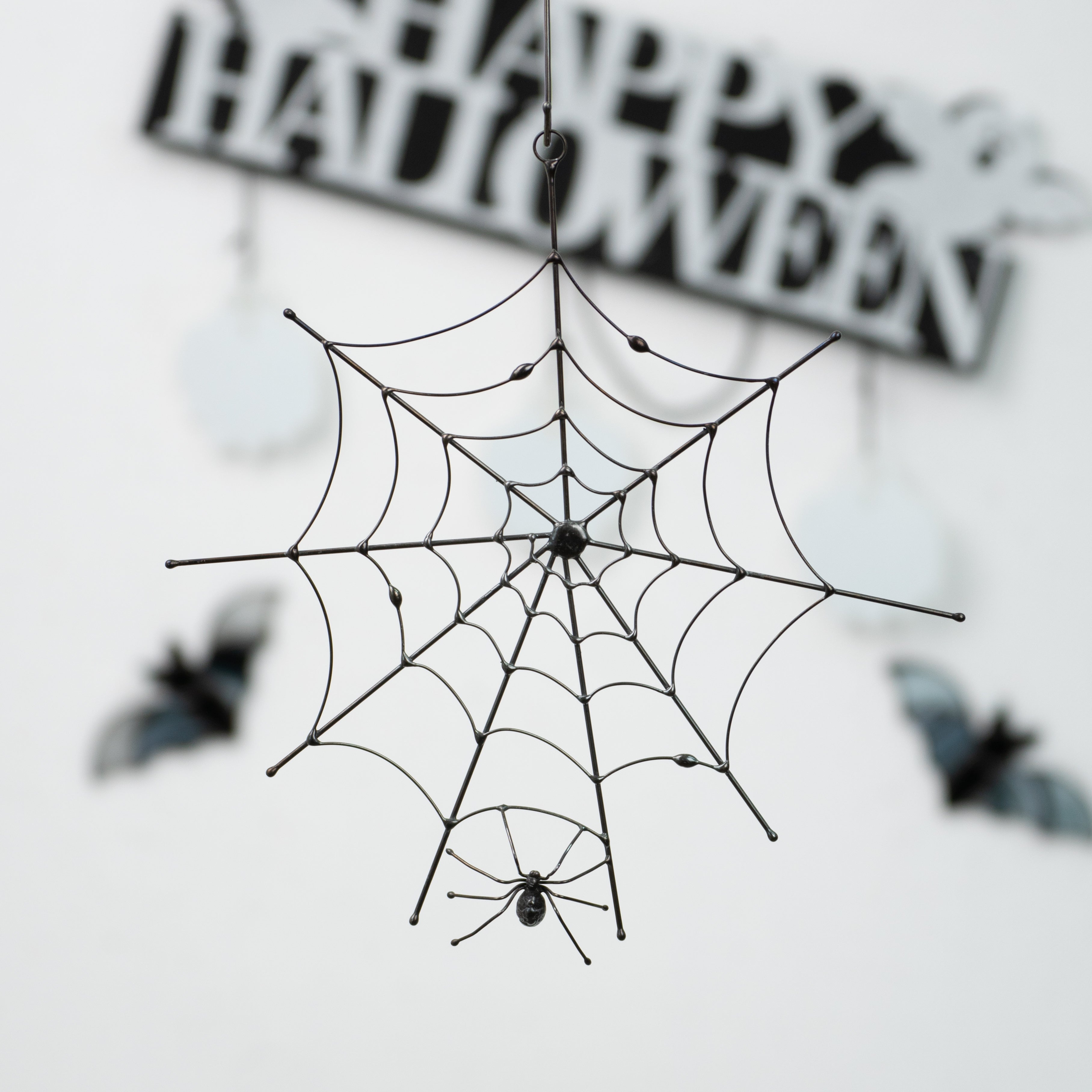 Metal spider web with a hanging spider, displayed in front of Halloween-themed wall decor with bats and a “Happy Halloween” sign.
