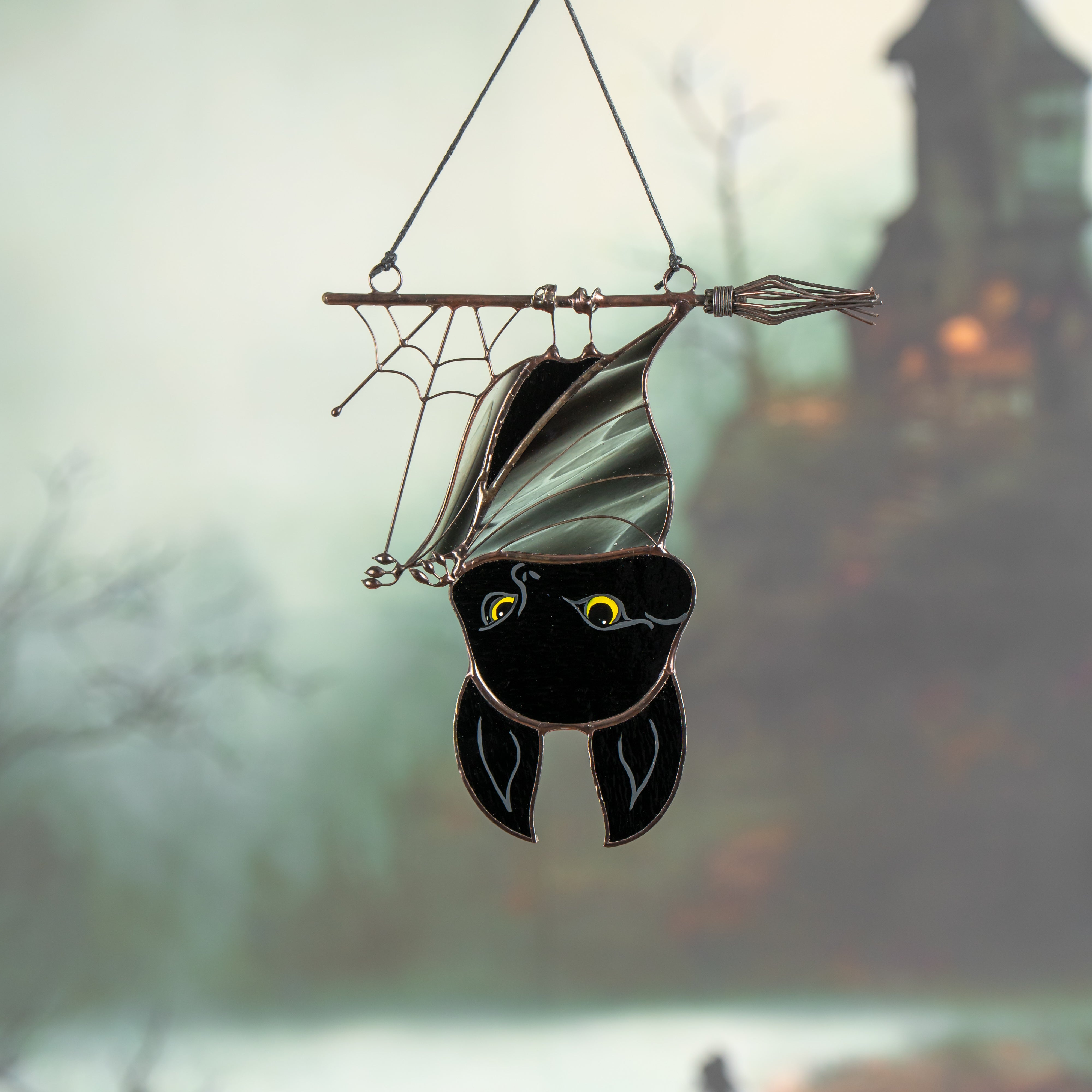 Black stained glass bat on broomstick shown with foggy haunted house background, glowing yellow eyes.