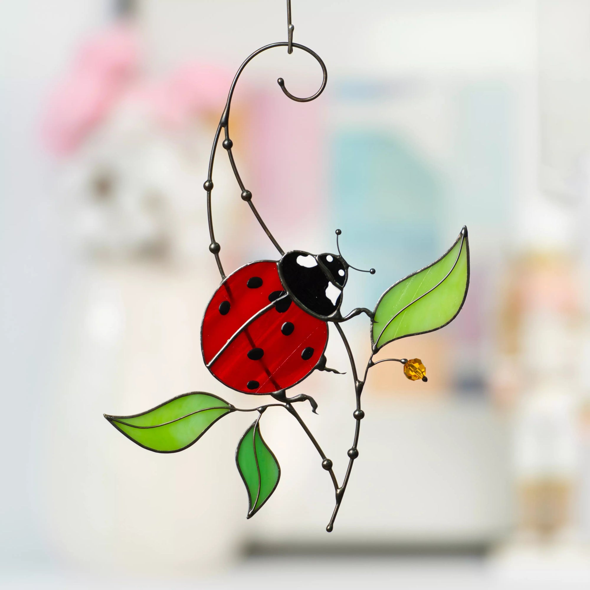 Ladybug stained glass with red wings, black spots, and green leaves hanging delicately on a metal wire with a leafy design.