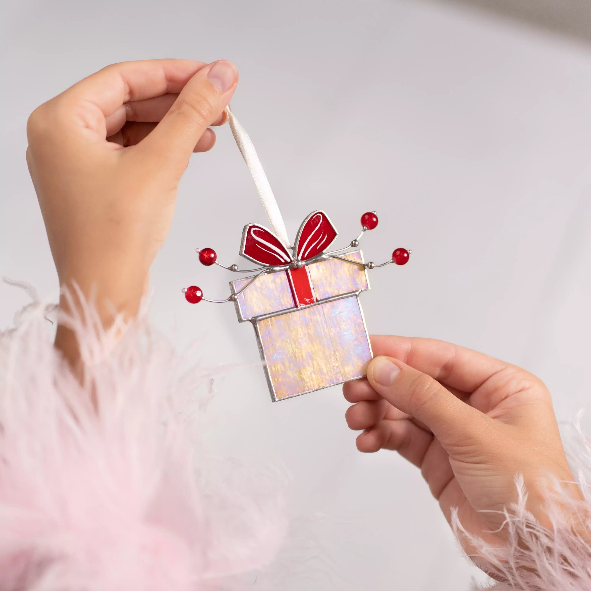 Hands holding stained glass suncatcher Christmas gift box ornament with red bow and festive red beads.