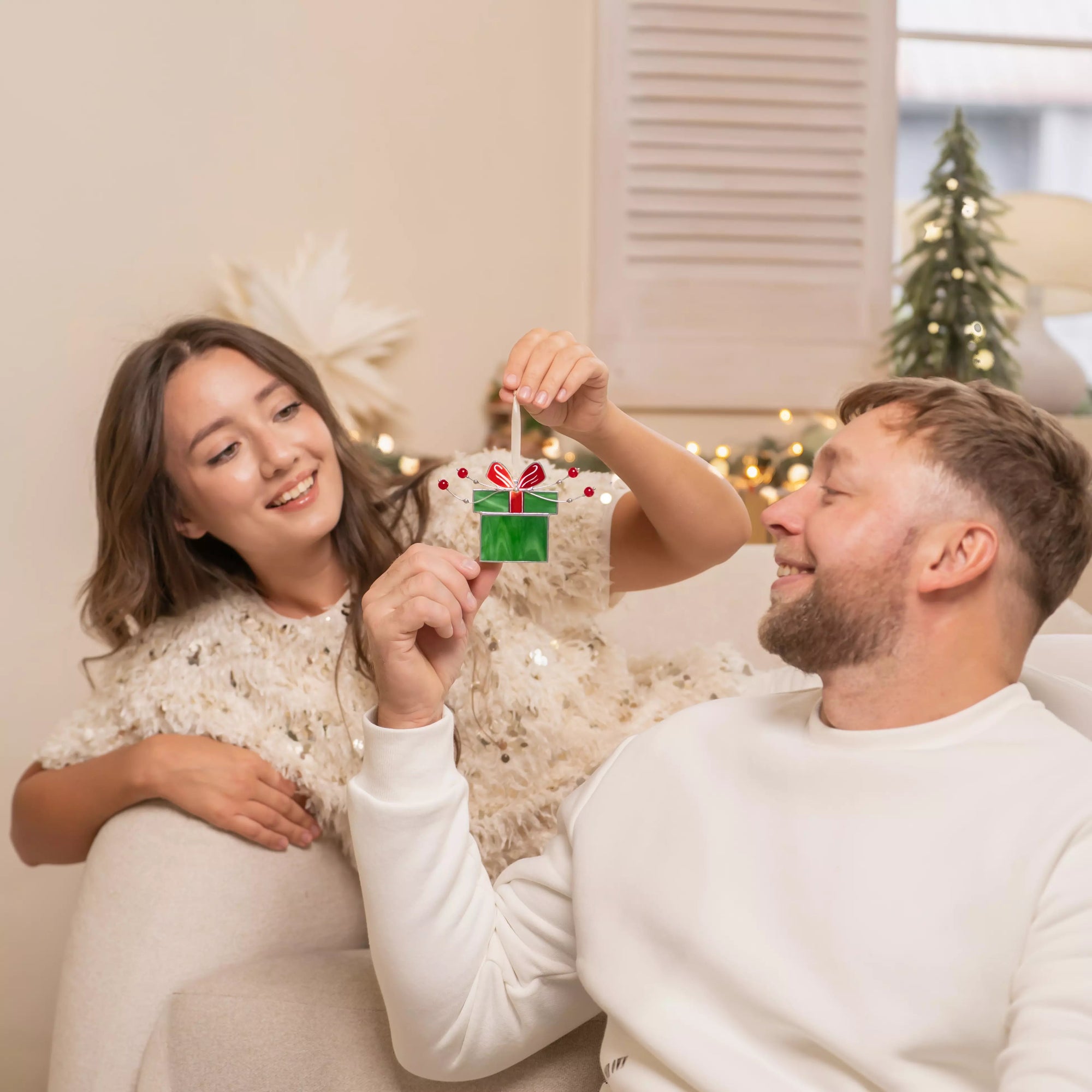 Two persons holding a stained glass green gift box with a red ribbon, adorned with small red beads and silver wire.