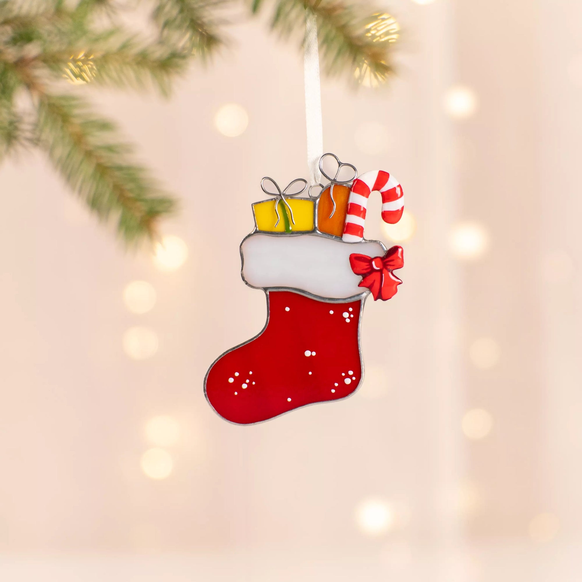 Red stained glass Christmas stocking suncatcher with white trim, yellow and orange presents, and a striped candy cane hanging on a ribbon near pine branch with warm lights.