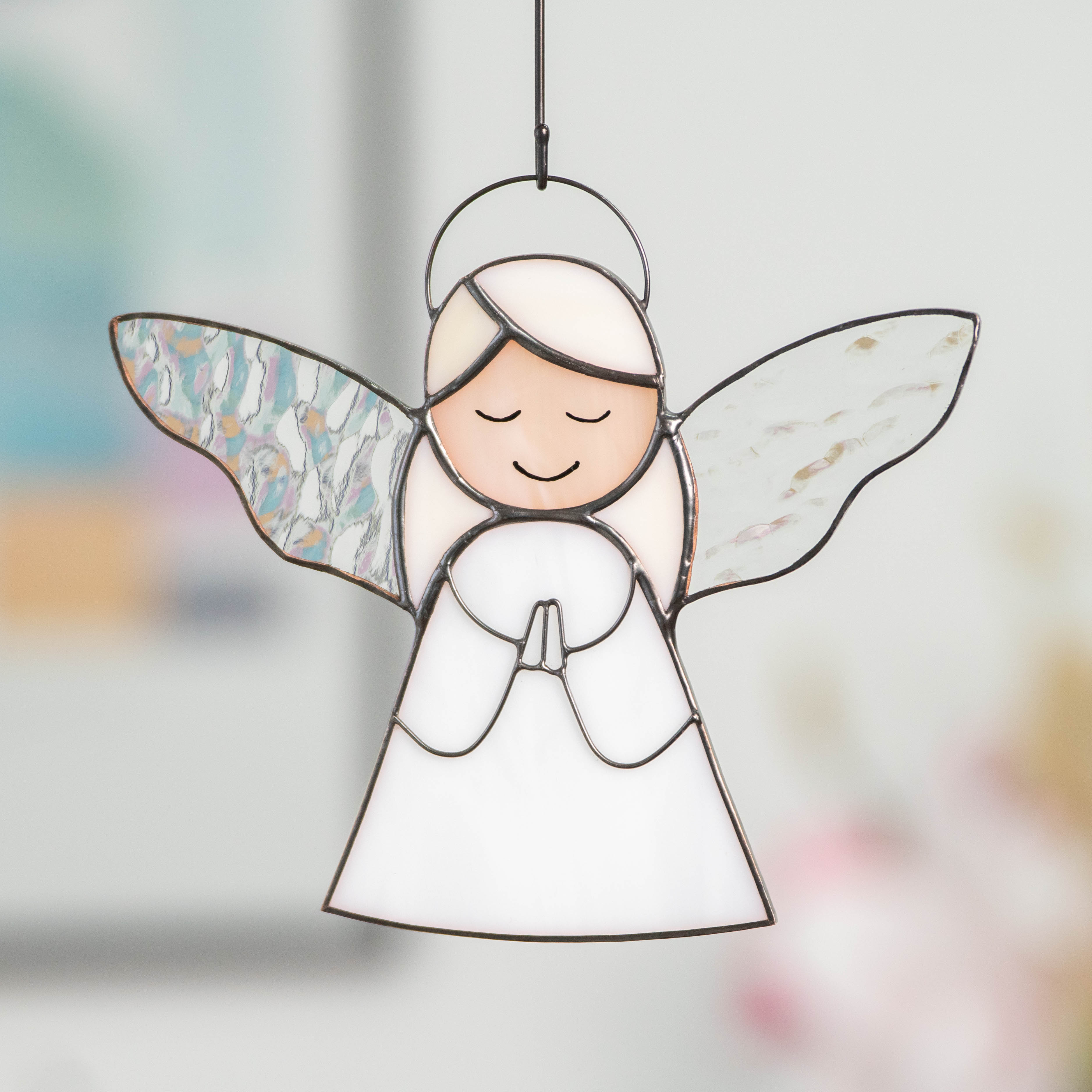 Handcrafted stained glass angel with closed eyes and praying hands, hanging in a softly lit interior background.