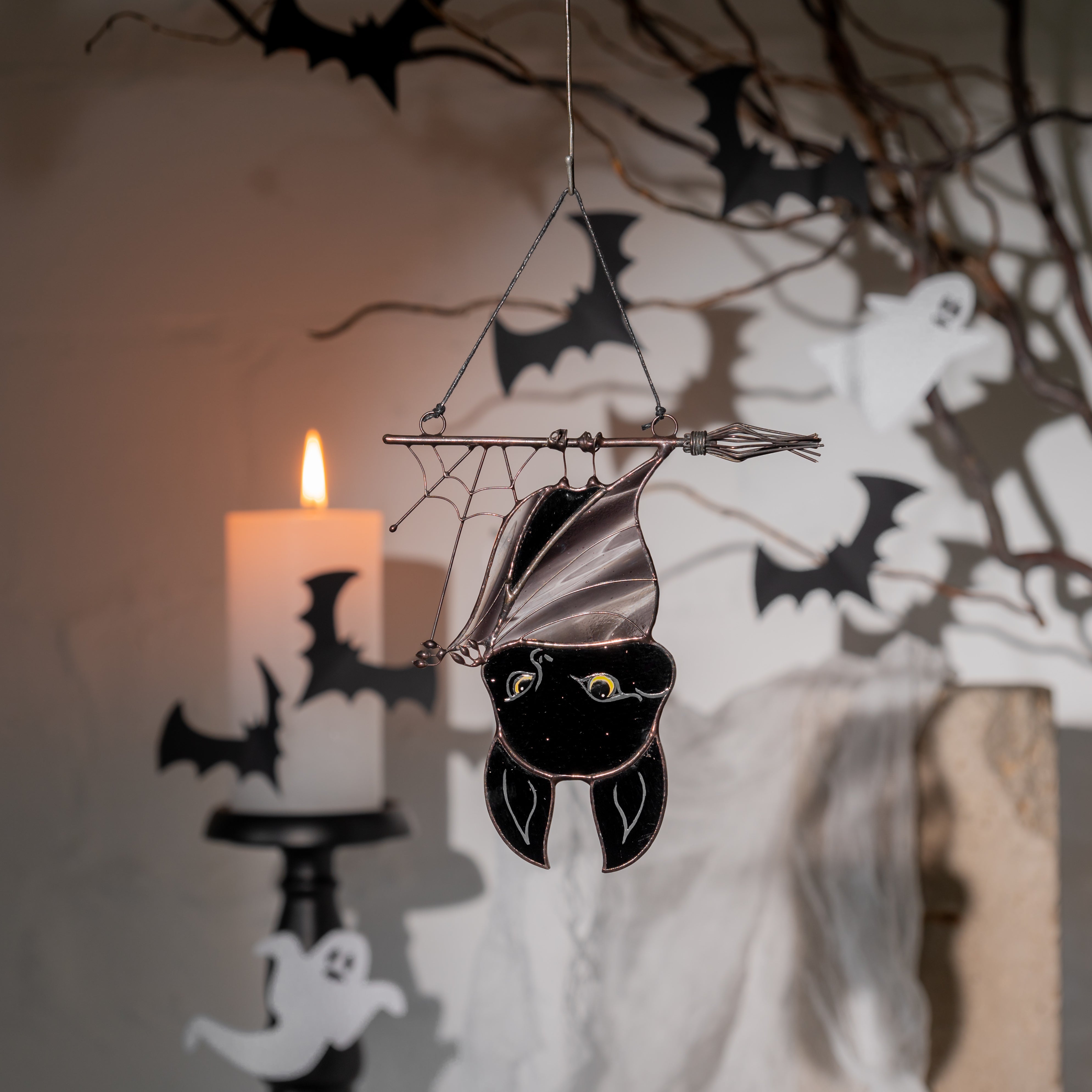 Cute bat stained glass hanging among Halloween decor with candles, bats, and ghosts in the background.